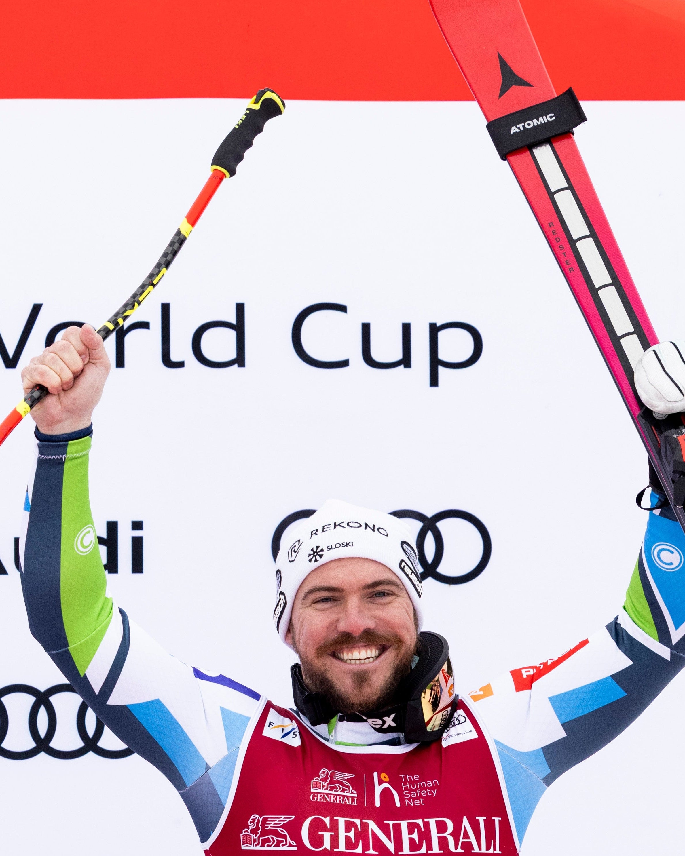 Miha Hrobat in a red, white, and blue ski suit celebrates on a podium, holding skis and a ski pole in front of an Audi World Cup backdrop. The bib shows the number 21.