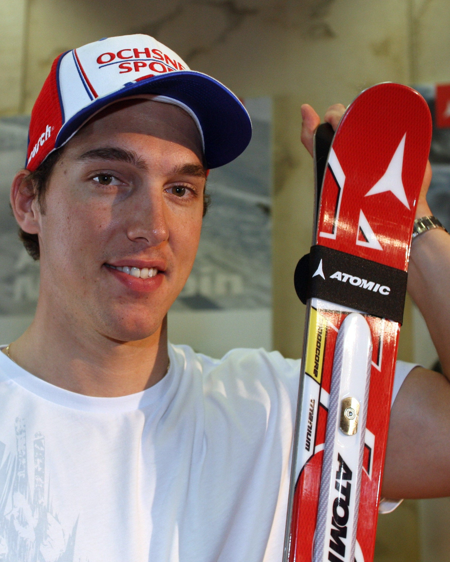A smiley Carlo Janko wearing a white t-shirt and a red, white, and blue cap holds up a pair of red and white Atomic skis with a black wrist strap in an indoor setting.