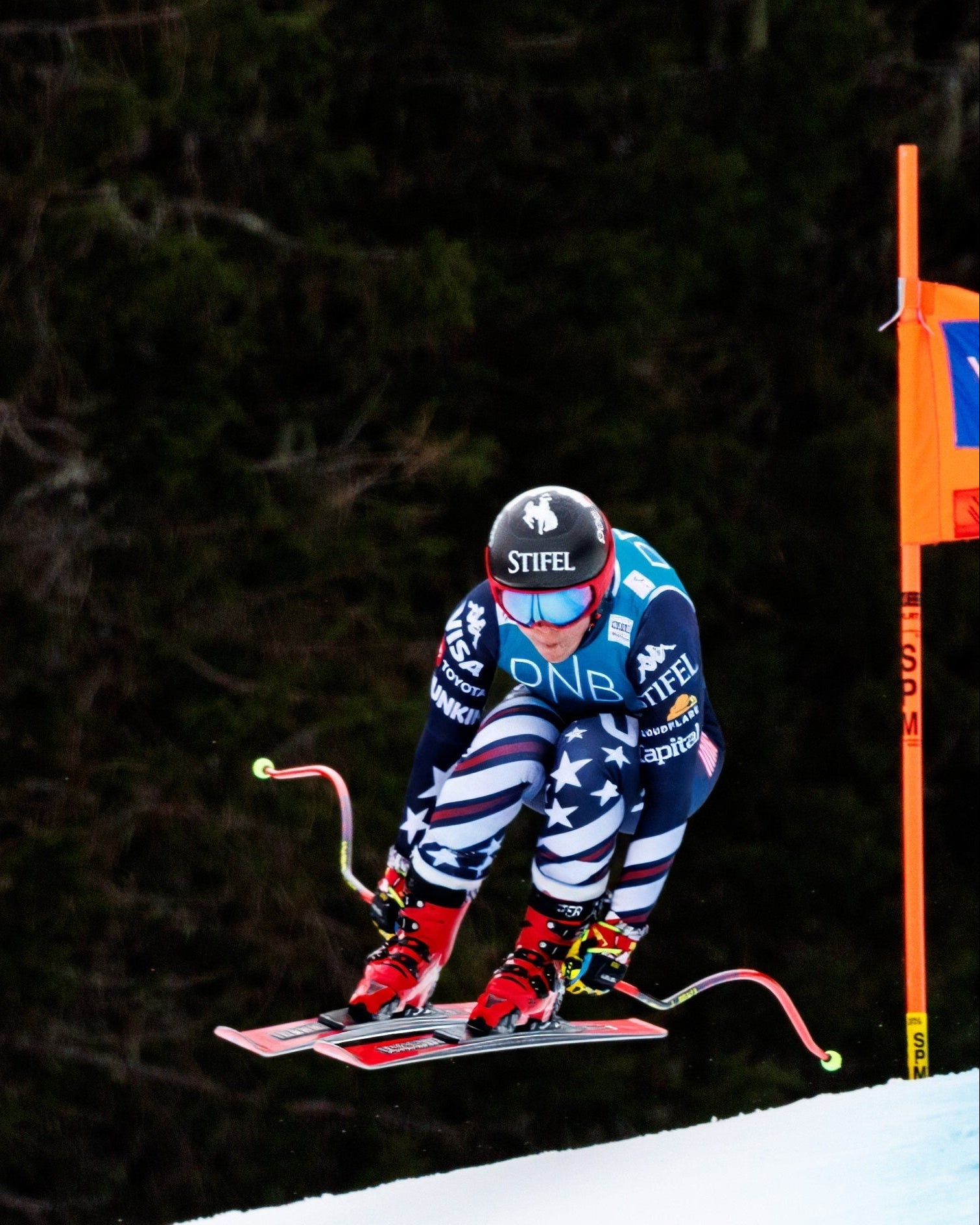 Breezy Johnson in a blue, red, and white patriotic patterned ski suit jumps through a gate on a snowy slope, with a dark forest background and an orange gate on the right.