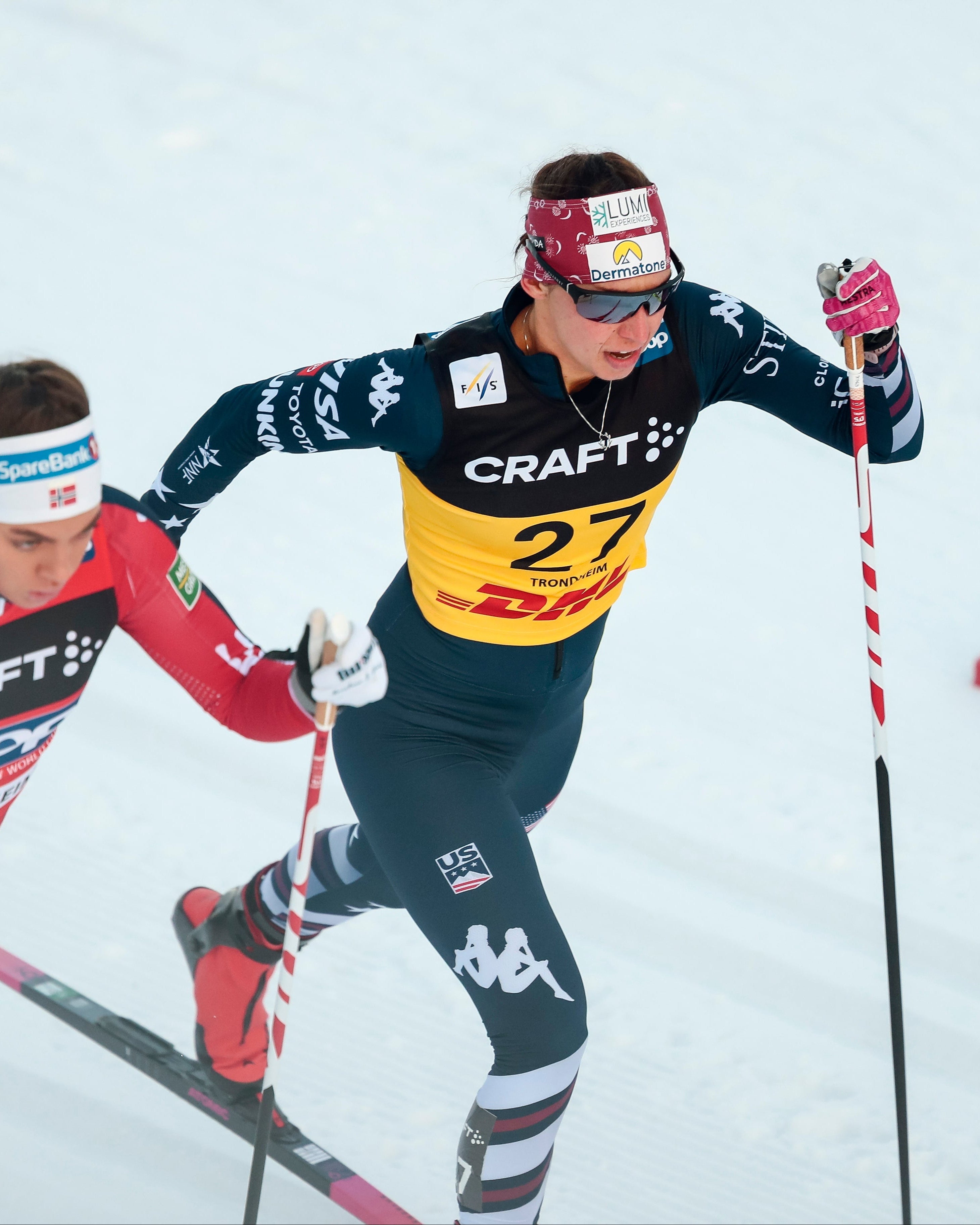 Julia Kern in a dark navy and yellow ski suit, wearing a red headband and goggles, racing on a snowy track alongside another racer in a red suit.
