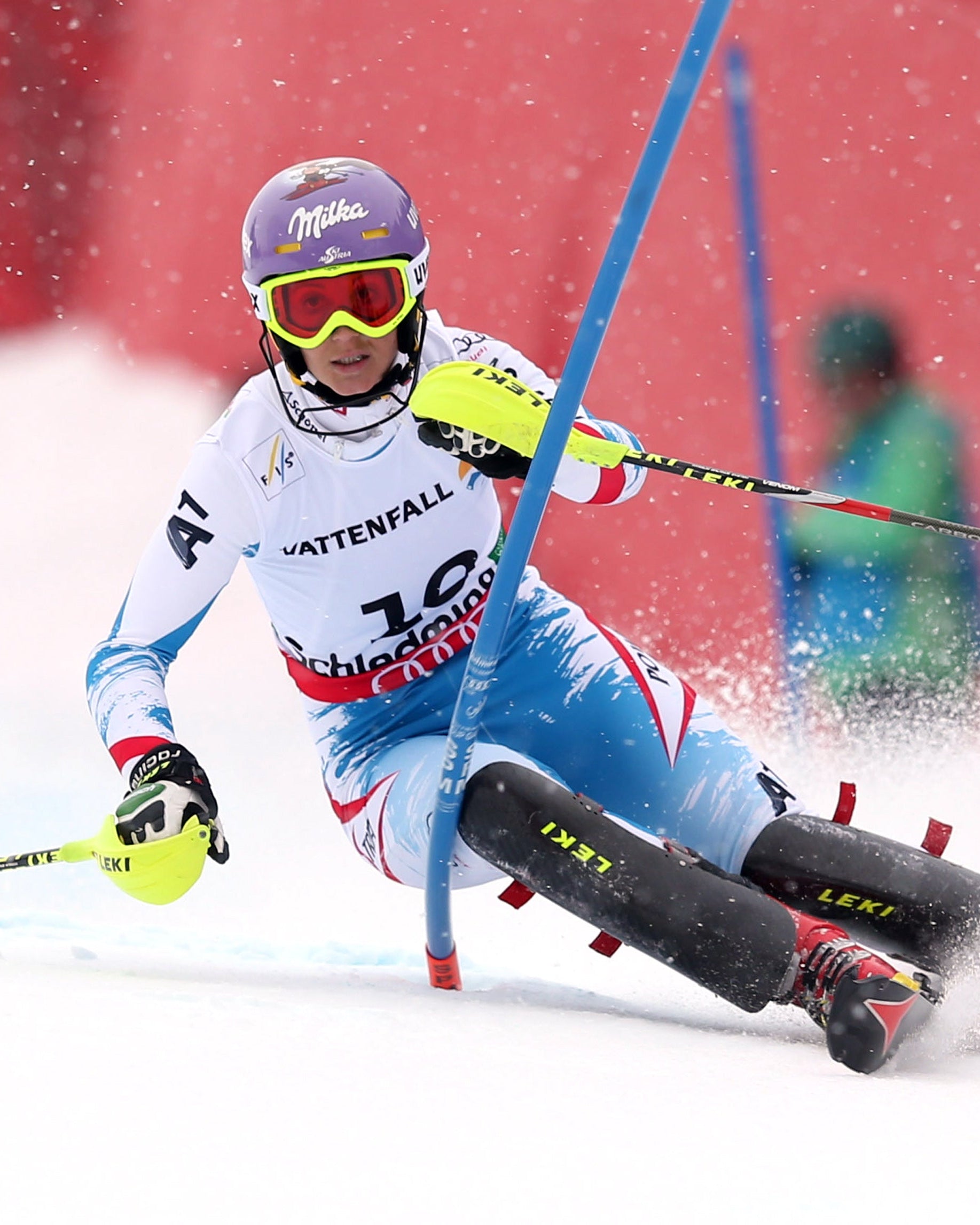 Michaela Kirchgasser in a white and light blue racing suit with 'Mattenfall' and 'Schwabenalp' branding, wearing a purple helmet and goggles, carving through fresh snow with ski poles and blue gate poles visible.