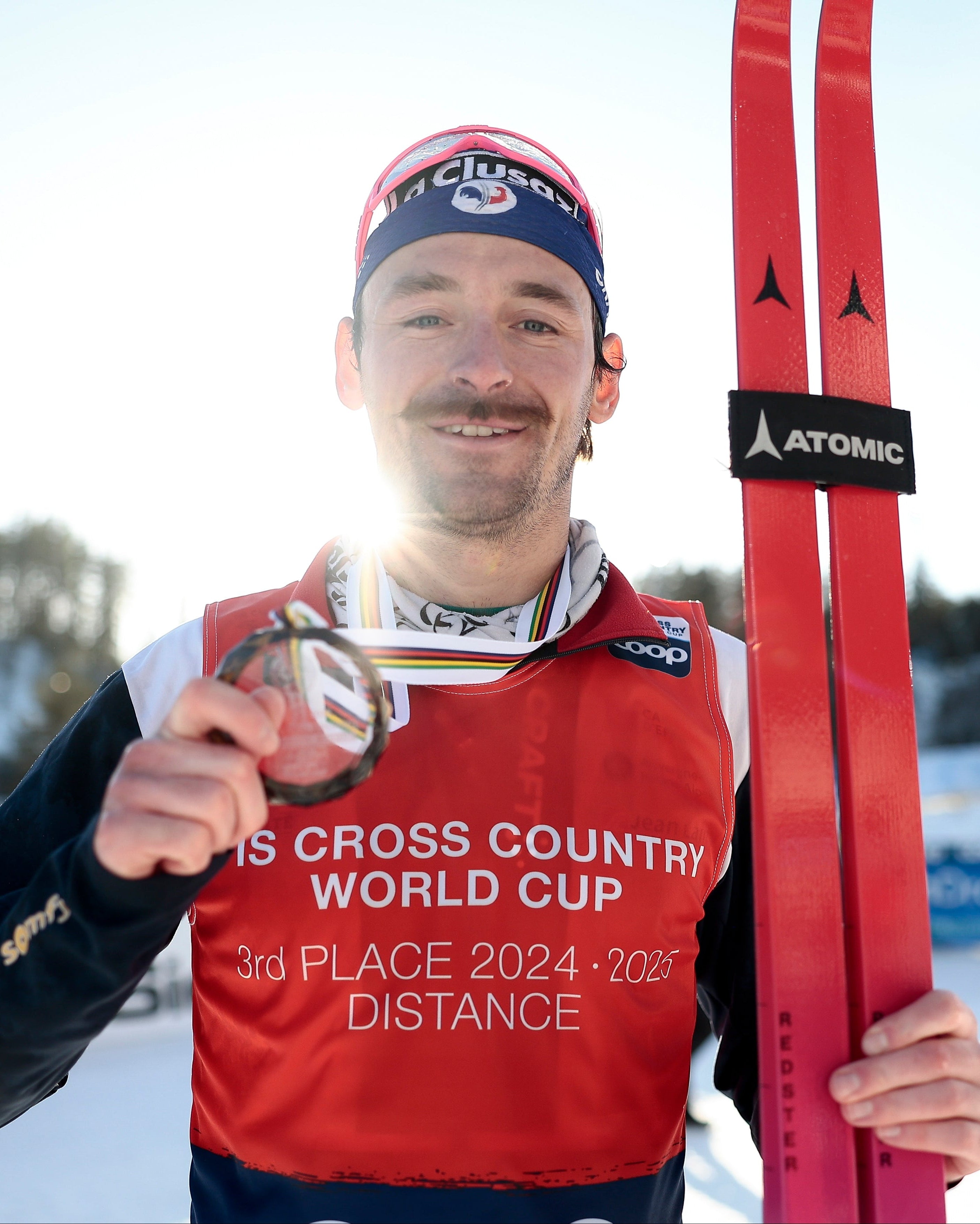 Hugo Lapalus in a red bib and black beanie holds up a medal while standing outdoors in the snow, holding red skis with visible ATOMIC branding.