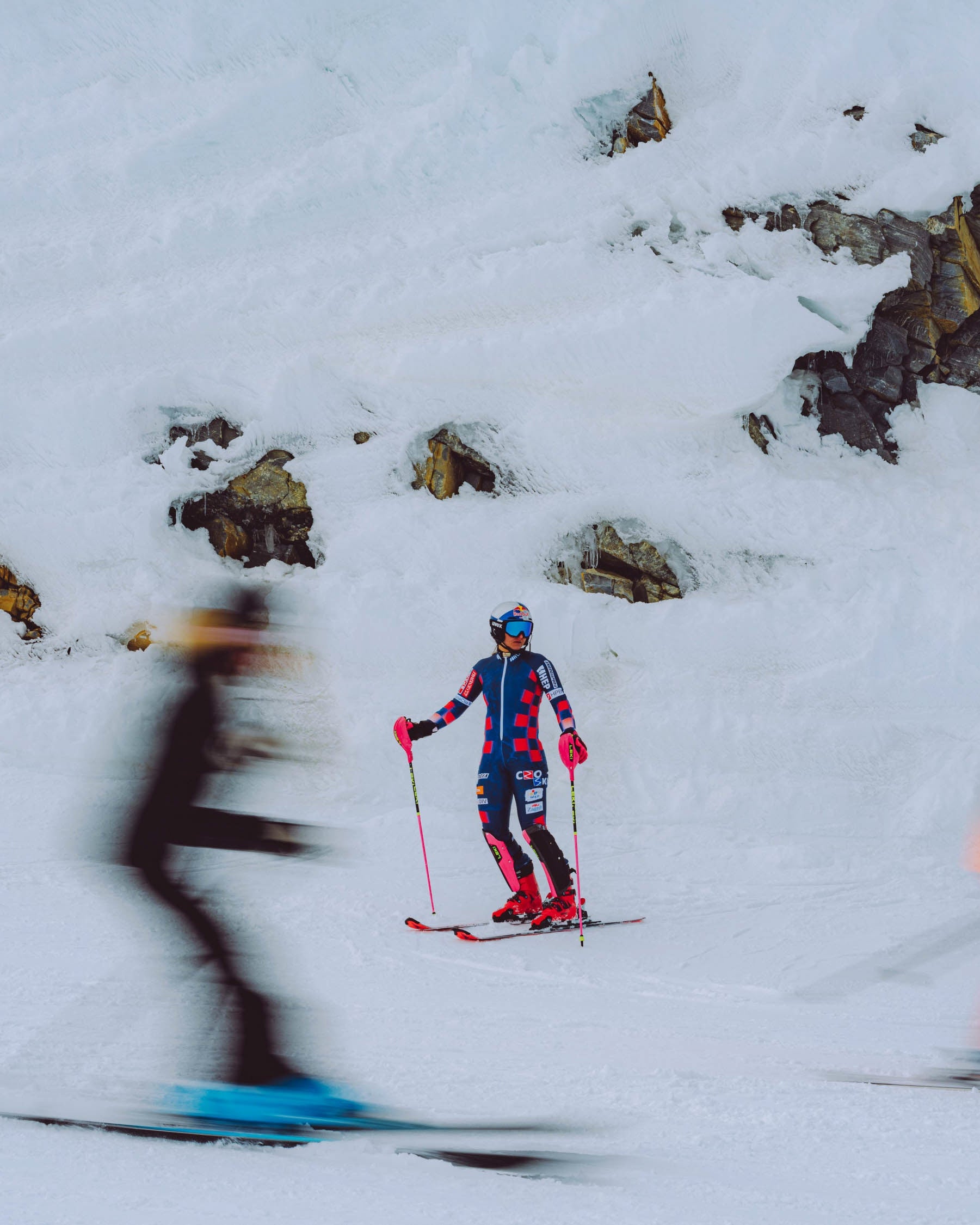 Zrinka Ljutic in a blue and red suit stands on a snowy mountain slope holding ski poles, with blurred skiers in the foreground and background and rocky snow patches behind them.