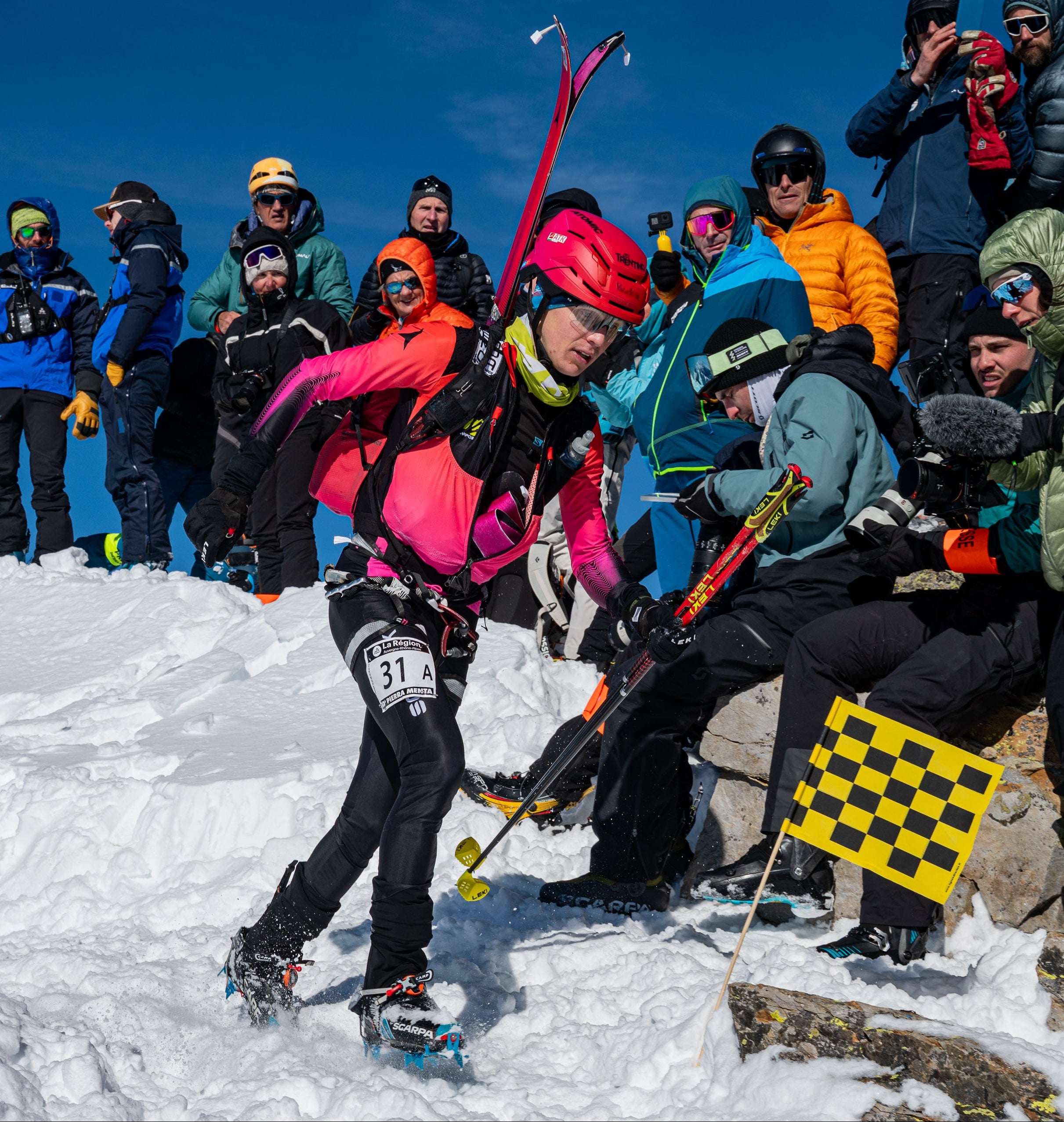 Davide Magnini in a bright pink jacket and red helmet navigates snowy terrain outdoors, carrying ski poles and skis, with spectators and other skiers in the background under a blue sky.