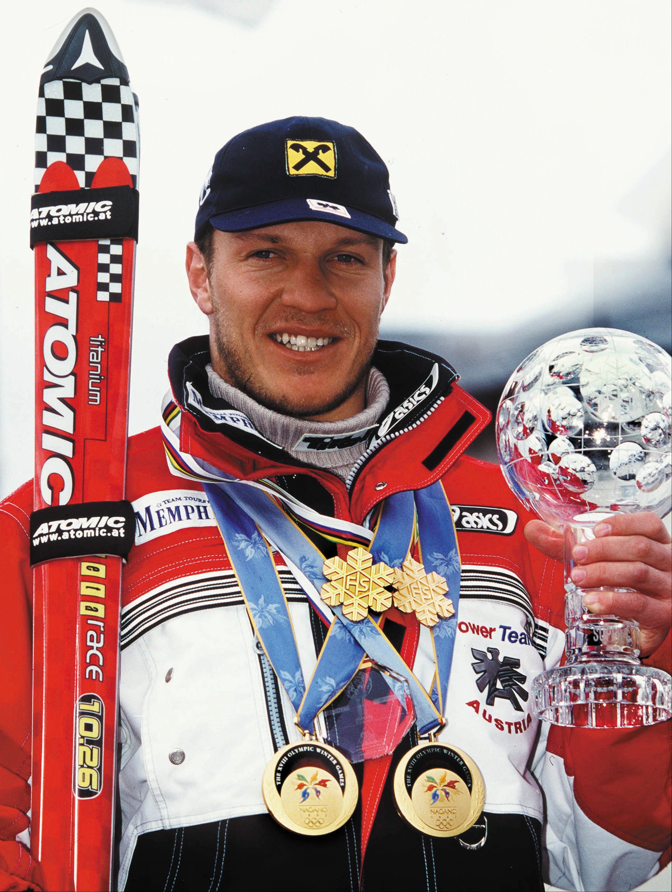 A smiley Hermann Maier in a red, white, and black ski racing jacket holds a crystal trophy while wearing multiple gold medals and a dark blue cap. A red Atomic skis stand upright behind him against a bright snowy outdoor background.