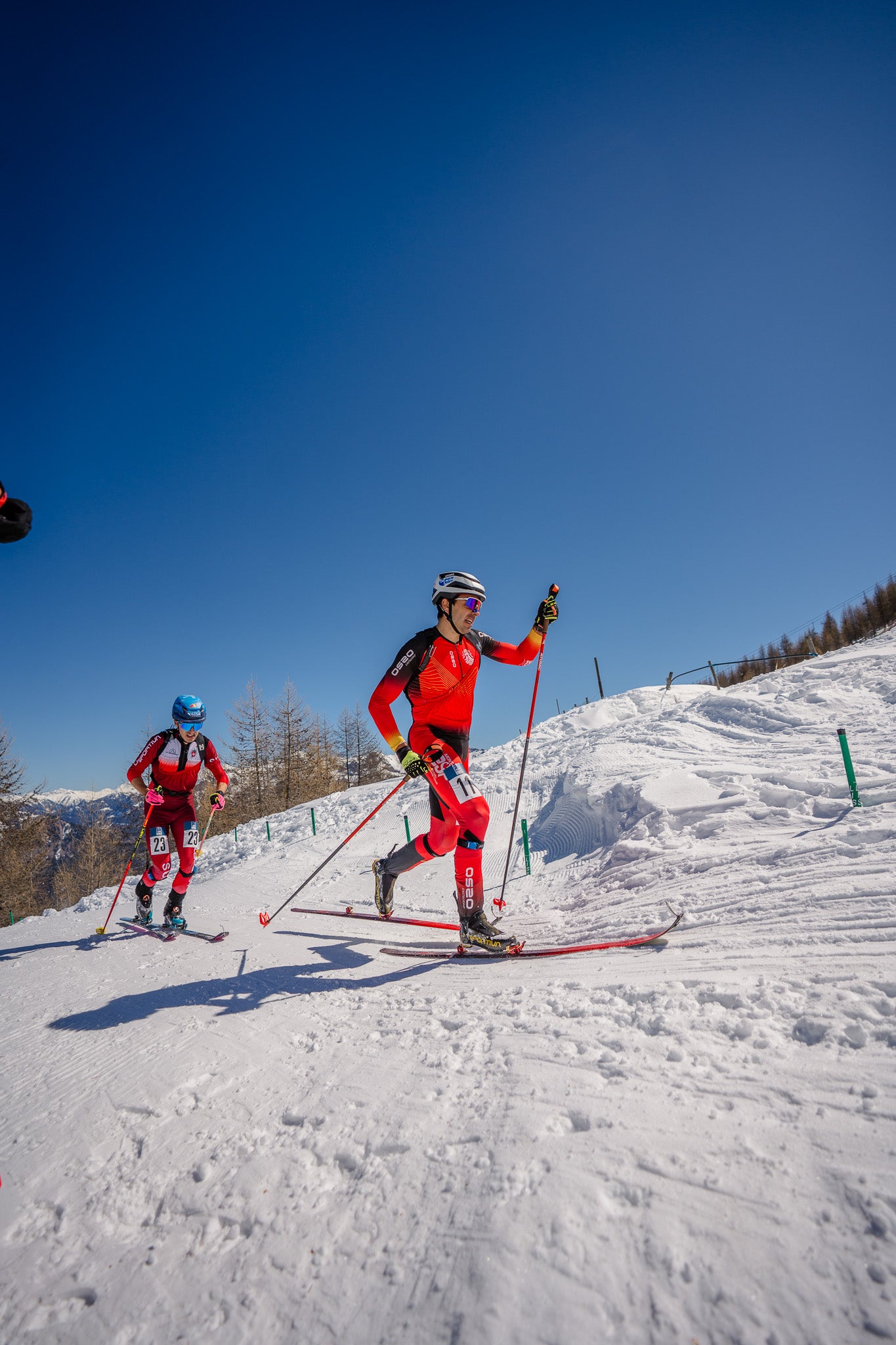 Two cross-country skiers racing up a snowy slope under a clear blue sky. The skier in the foreground wears a bright red and black ski suit with red pants and holds ski poles, while another skier follows behind in a red suit with a blue helmet.