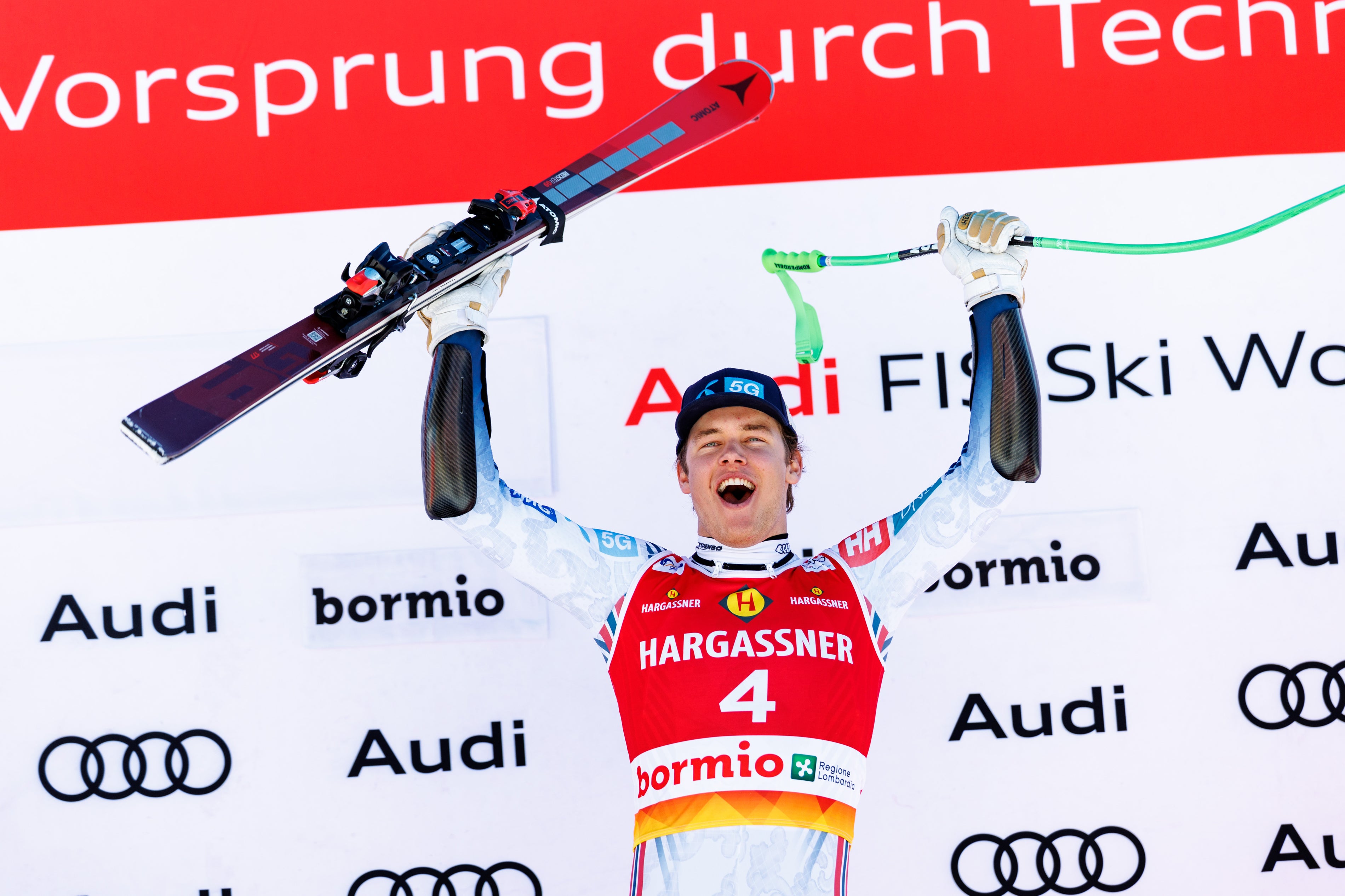 Fredrik Moller celebrates on a podium with arms raised, holding skis above his head. He wears a red, white, and blue Hargassner ski racing jersey with Audi and bormio sponsor logos.