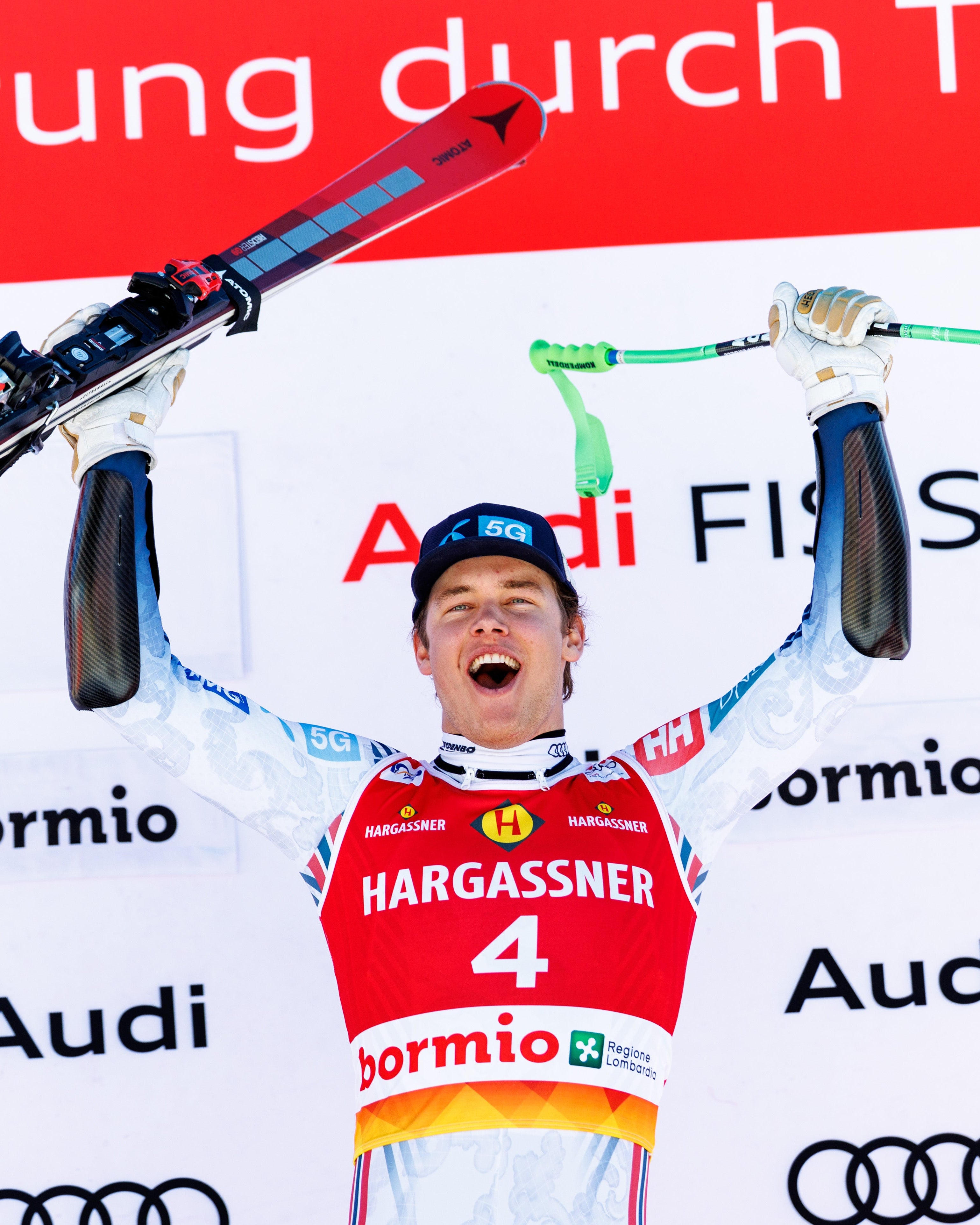 Fredrik Moller celebrates on a podium with arms raised, holding skis above his head. He wears a red, white, and blue Hargassner ski racing jersey with Audi and bormio sponsor logos.