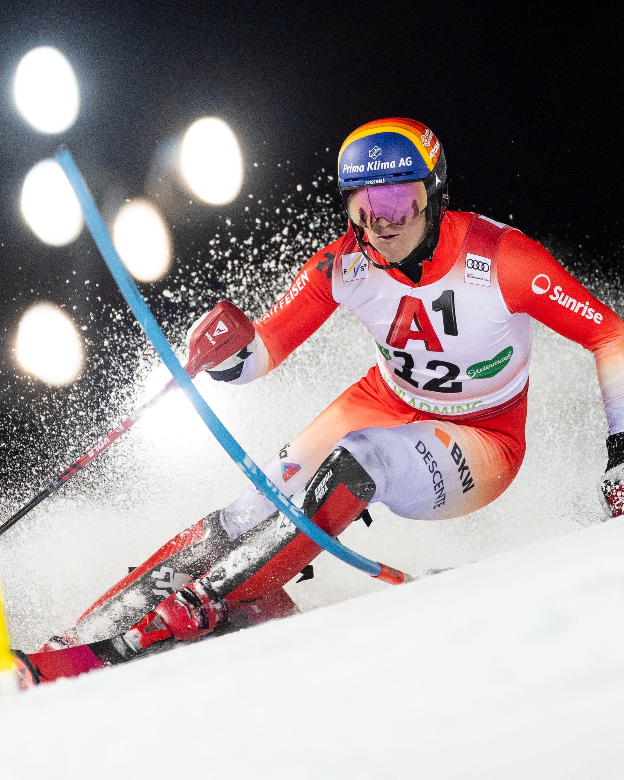 Tanguy Nef in a red and white racing suit with the number 12 and sponsor logos, carving through snow at night under bright stadium lights.