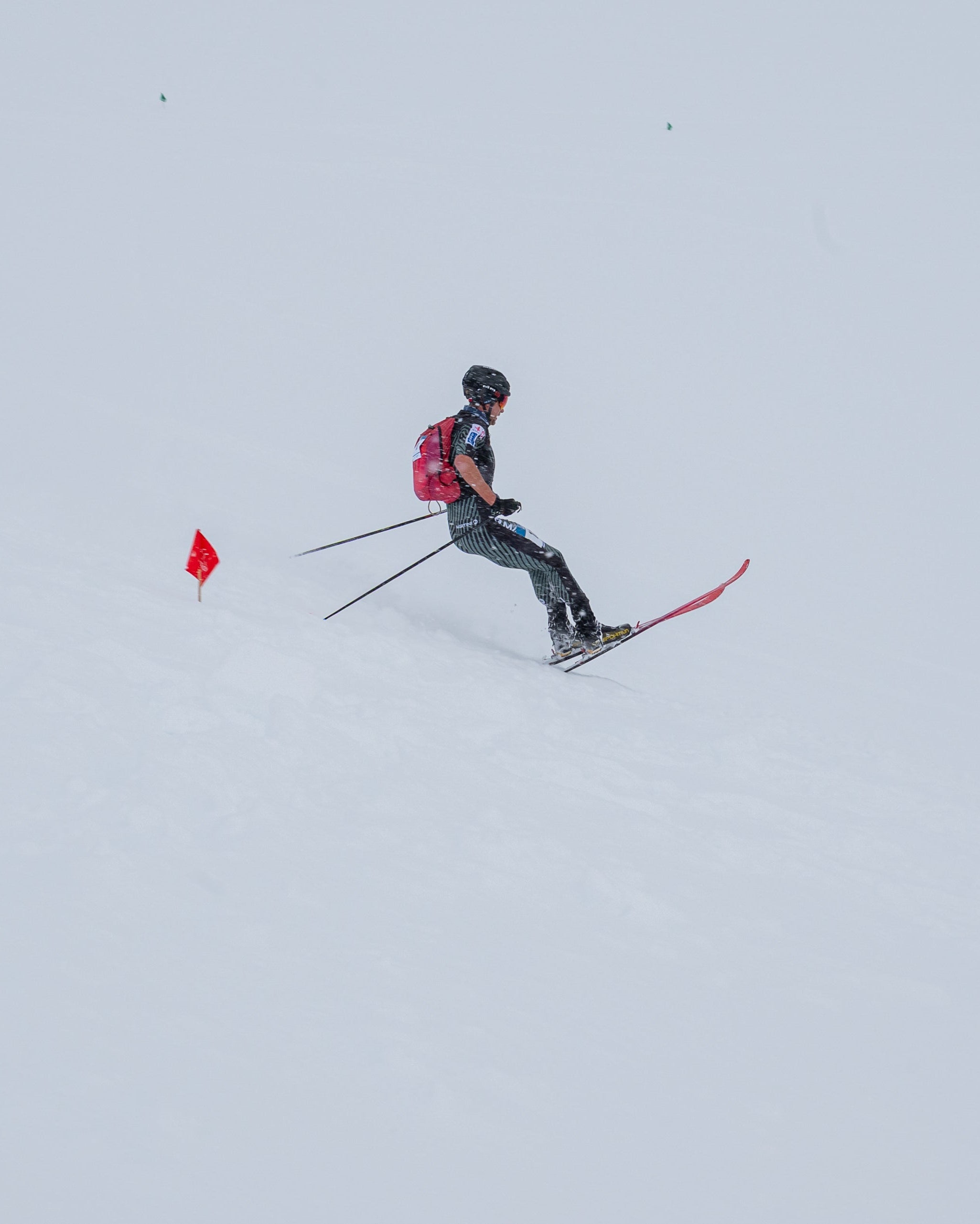 Nils Oberauer in dark clothing with a red backpack and red skis moves across a snowy slope, with a small red flag from the race course is visible to the left.