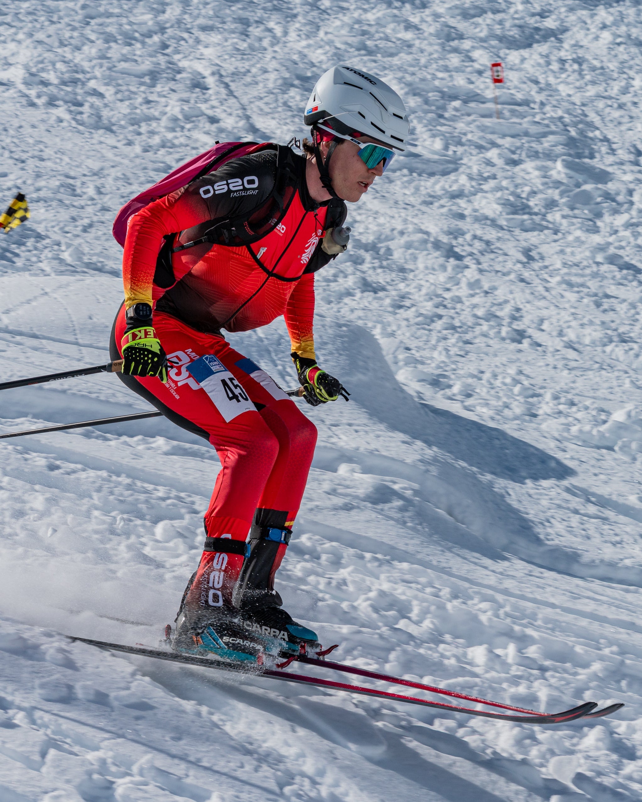 Albert Pérez in a red and black ski suit with a white helmet and sunglasses carving through fresh powder snow, holding ski poles.