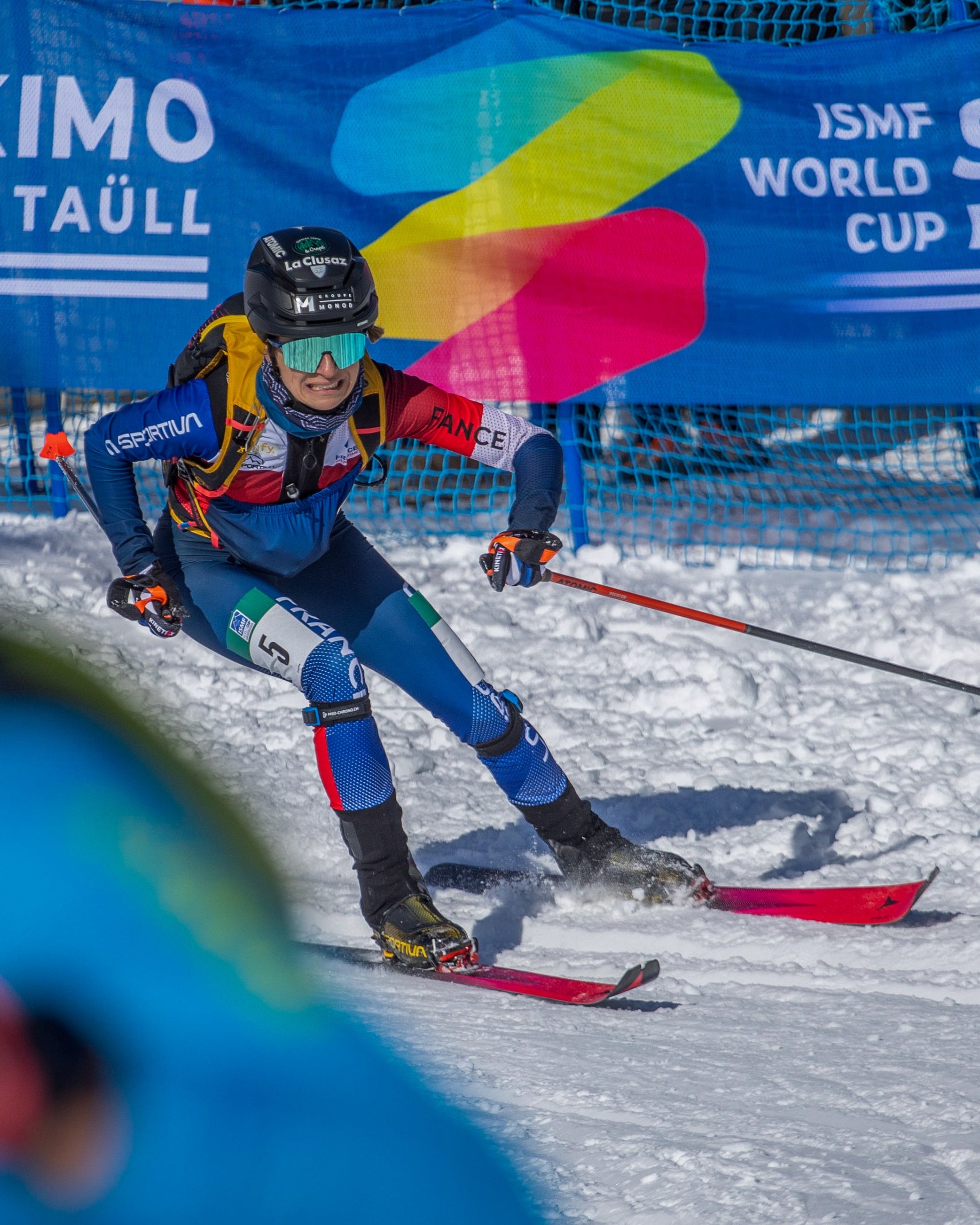 Celia Passey in a blue, red, and black racing suit navigates a snowy course with red skis, with a blue JSMF World Cup Bói Taúul banner in the background.