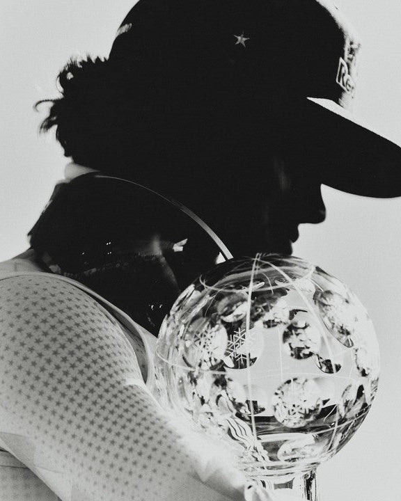 Black and white close-up of Lucas Pinheiro Braathen in profile wearing a cap and holding a large faceted crystal trophy against a light background.