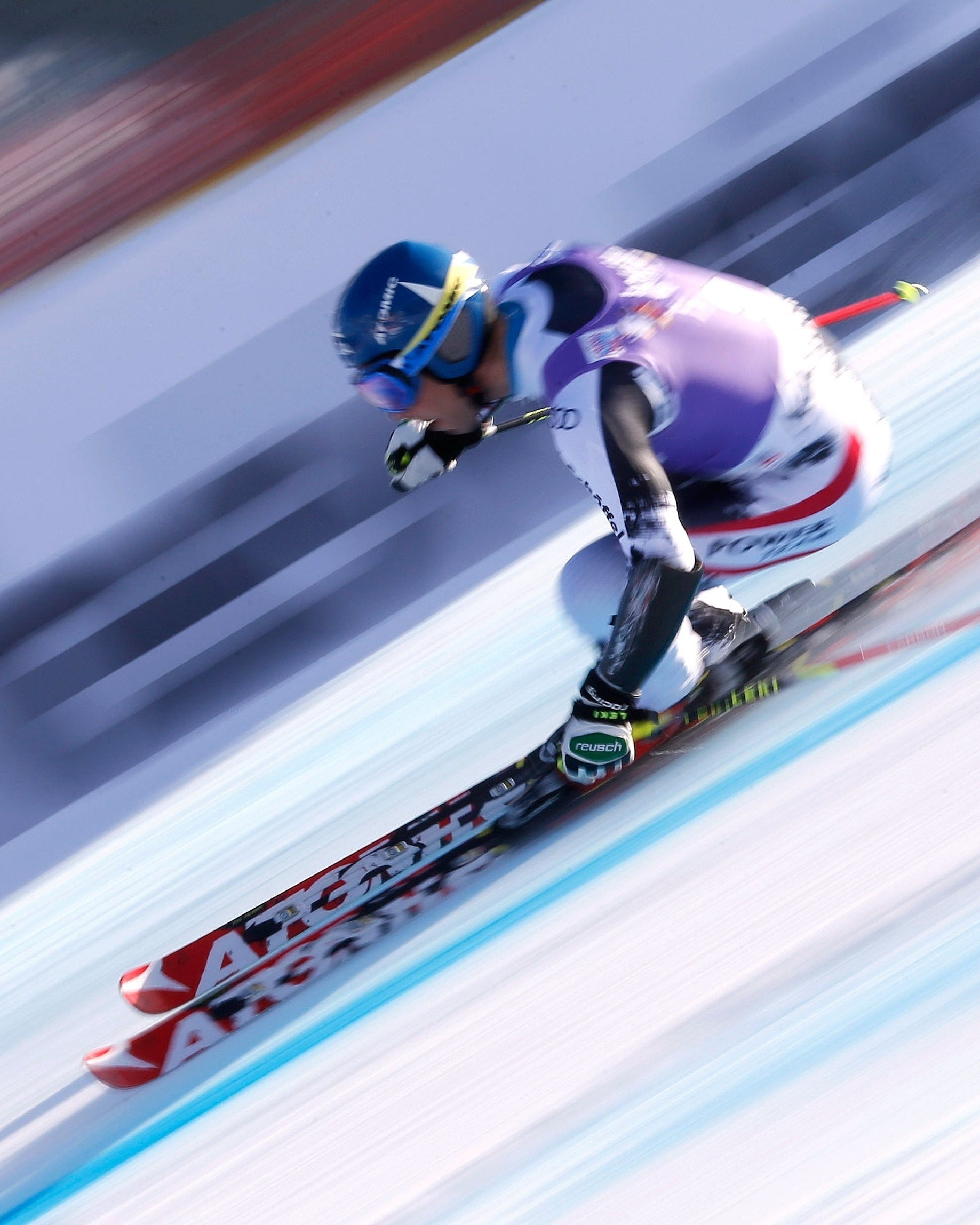 Benjamin Raich a white, purple, and black racing suit leans sharply into a turn on a snowy slope, with red and black skis and ski poles visible. The background shows a heavily blurred icy track and red-and-white barriers, emphasizing speed and motion.