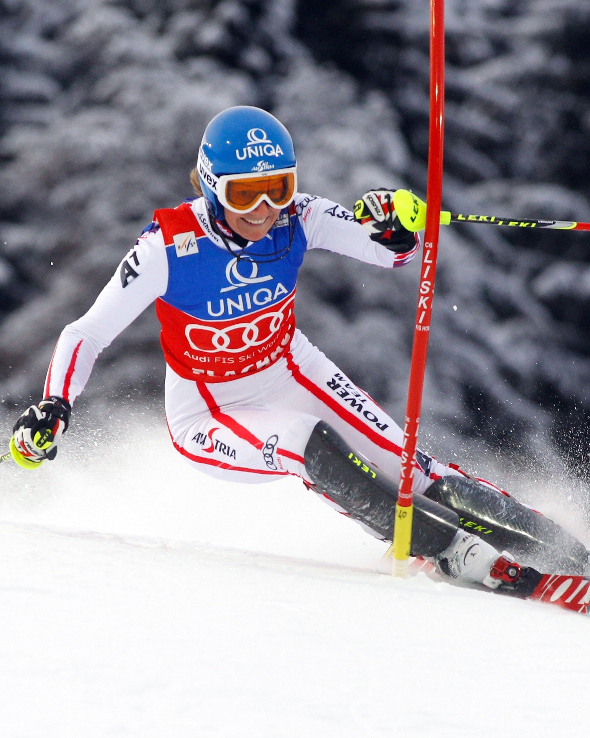 Marlies Raich in a white and red race suit leans sharply into a turn on snow, with a yellow pole on the right and dark, snow-covered trees in the background.