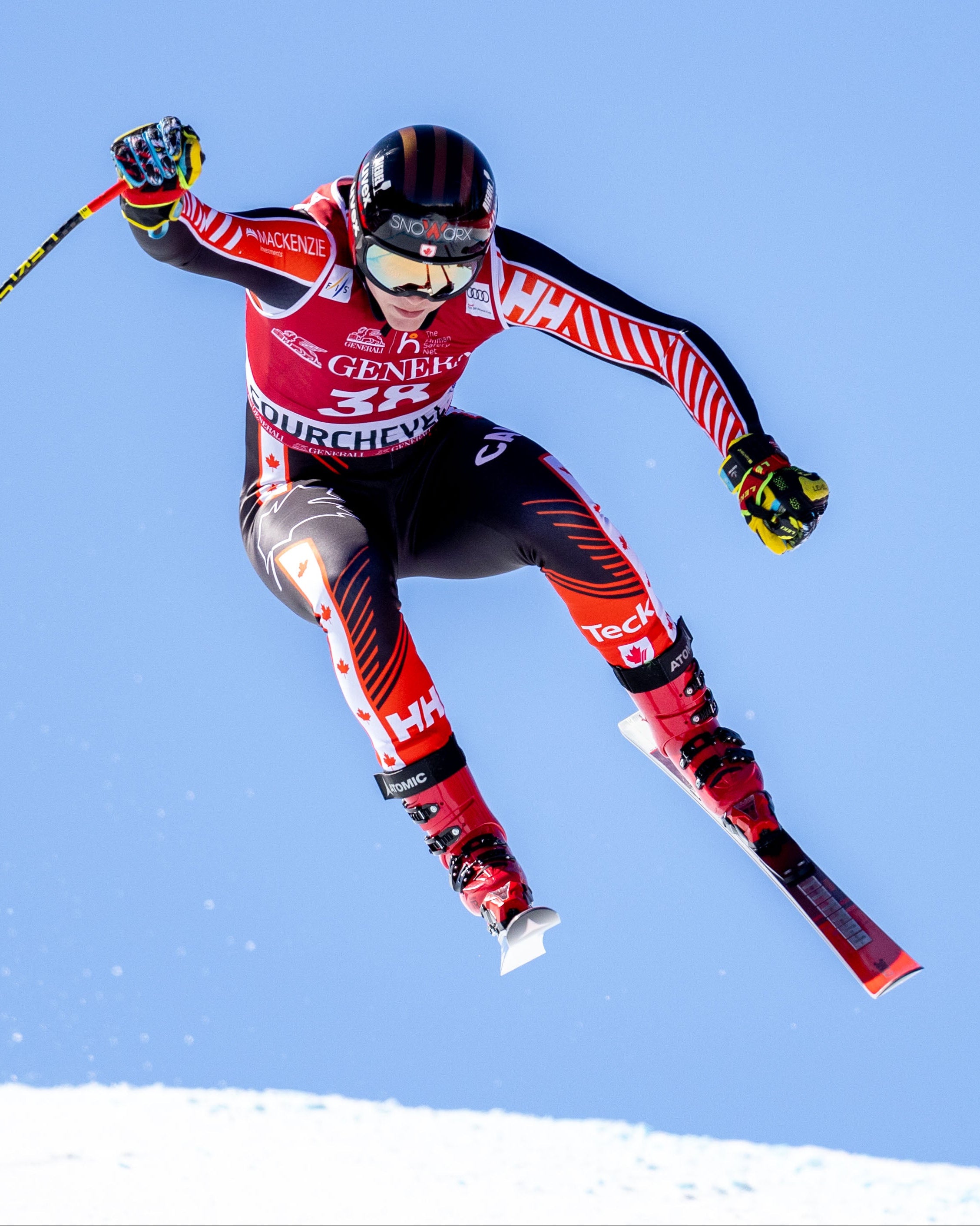 Jeffrey Read in a red, black, and white racing suit jumps through the air against a clear blue sky, holding ski poles.