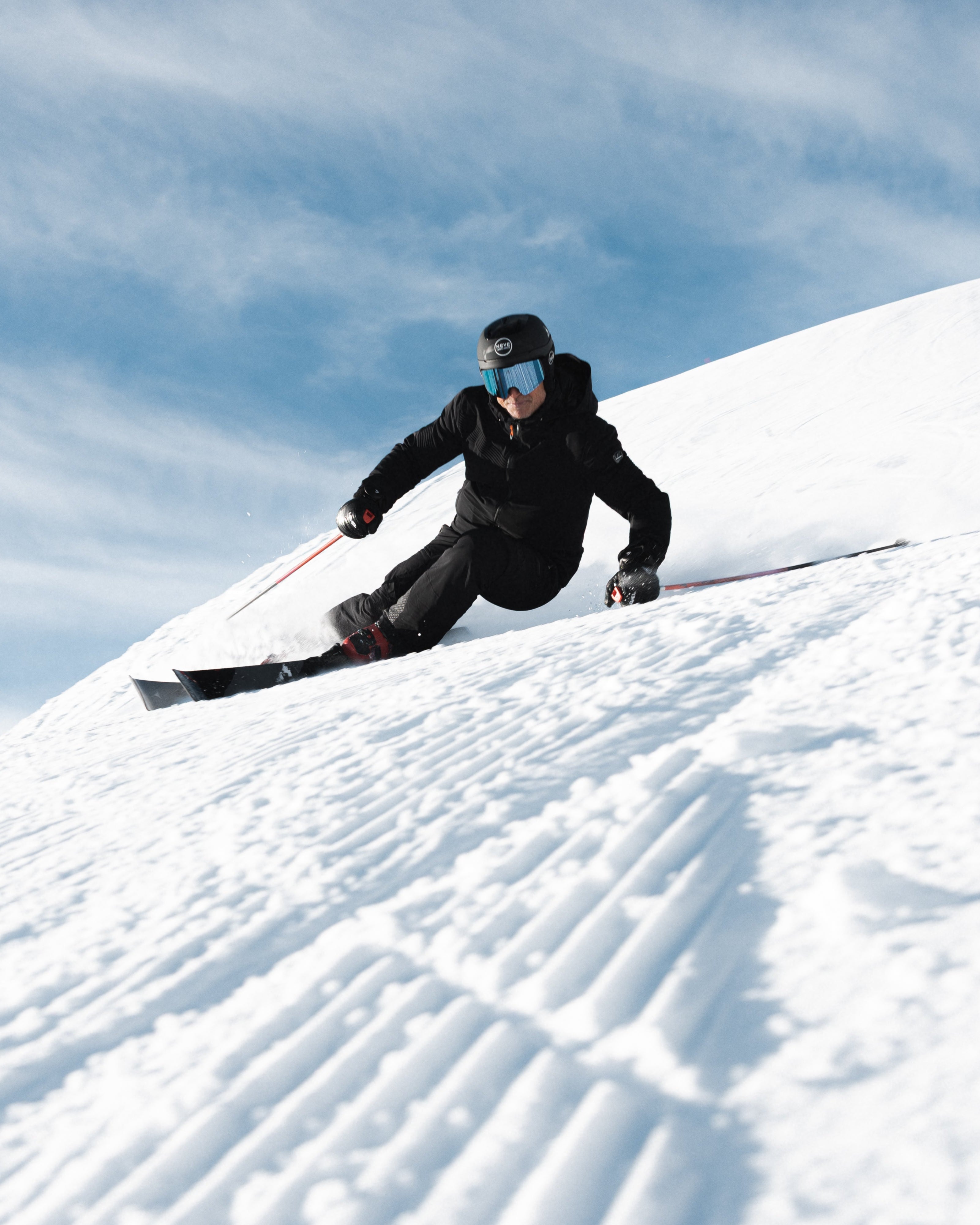 Giorgio Rocca in a black outfit and blue goggles slides down a snowy slope under a bright blue sky with wispy clouds.
