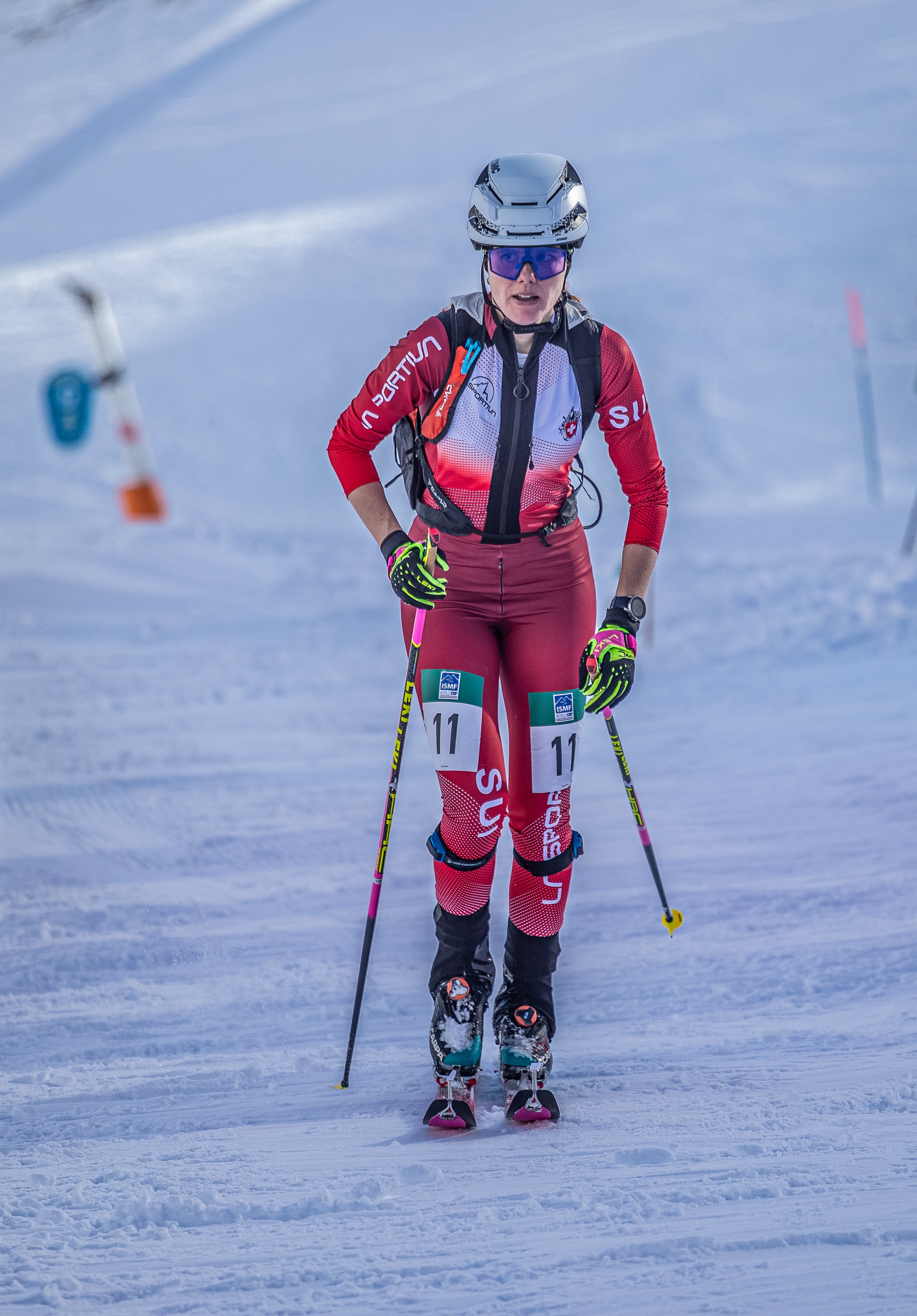 Alessandra Schmidt wearing a red, white, and black ski suit with bib number 11, a white helmet, and goggles, touring on a snowy slope with ski poles.
