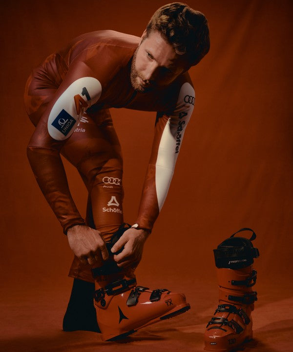 Marco Schwarz in a red racing suit adjusts his orange ski boot while another boot stands nearby on a warm brown studio background. 