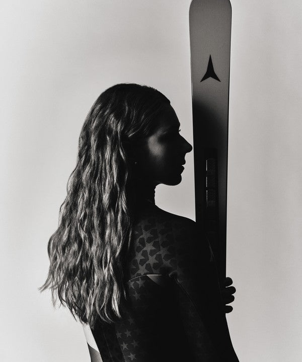 A black and white editorial photograph of Mikaela Shiffrin in profile holding a ski vertically next to her. The image is monochrome, highlighting the woman's long wavy hair, dark silhouette, and patterned race suit.