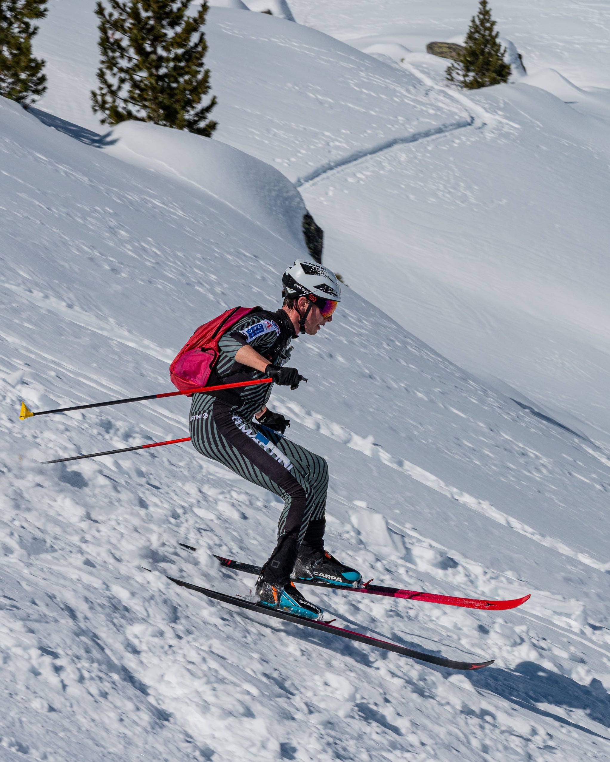 Julian Tritscher  in a black and gray racing suit with a red backpack and white helmet descends a snowy mountain slope, holding ski poles.