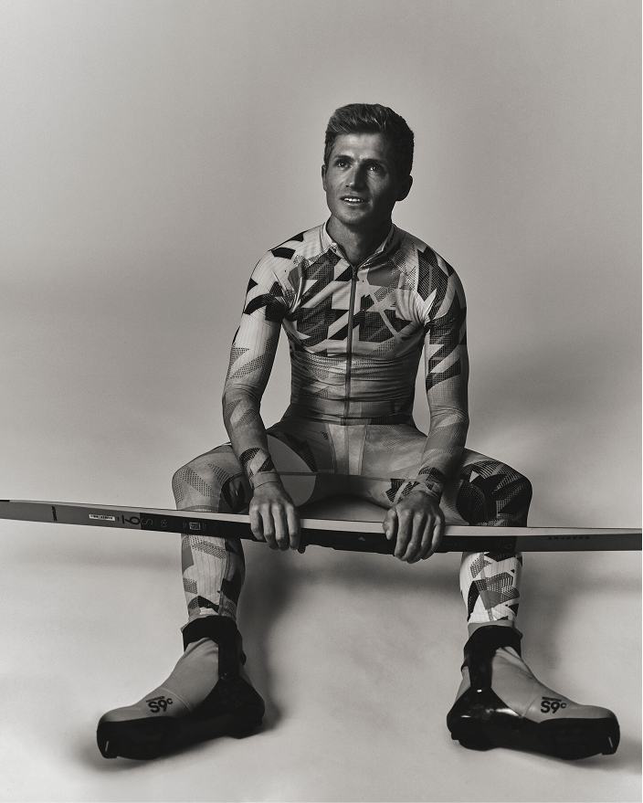 A black and white studio portrait of Mika Vermeulen sitting cross-legged and holding a long ski or snowboard, wearing a fitted geometric-patterned ski suit and ski boots against a plain light gray background.