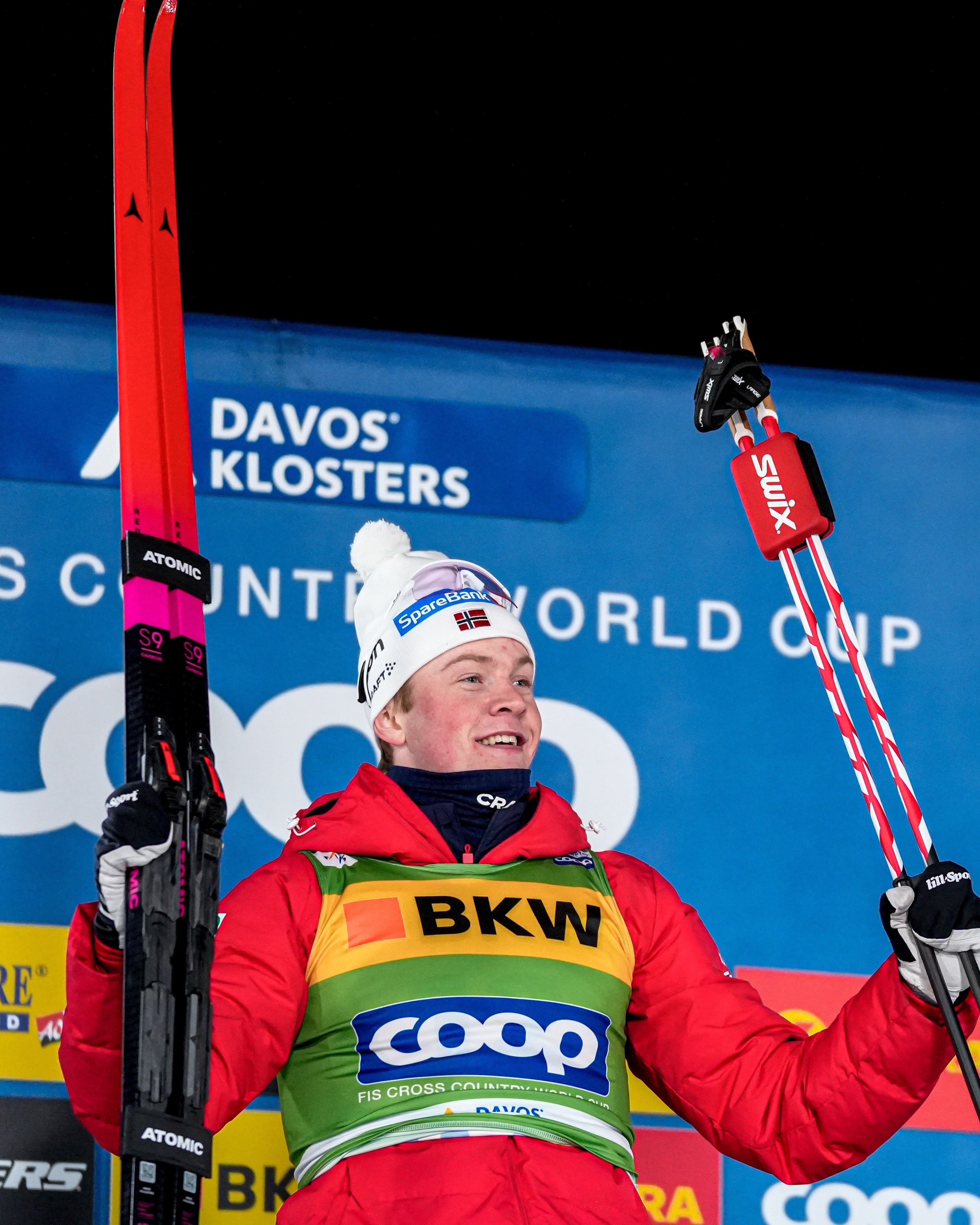A cross-country skier in a red, yellow, and green ski jumping suit holds red and black skis and poles, standing in front of a blue sponsor backdrop with logos including COOP, BKW, and Le Gruyère.