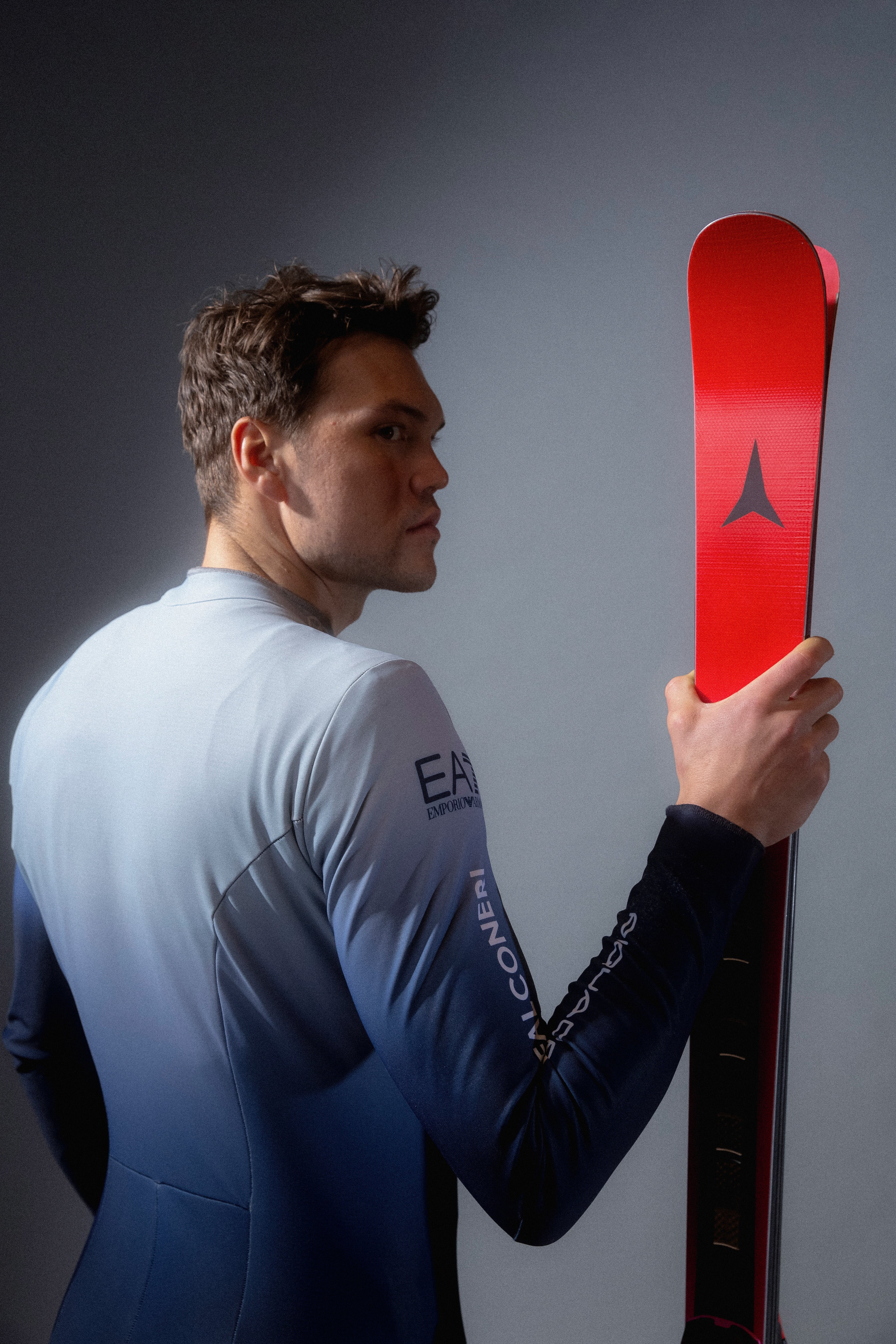 Aleks Vinatzer in profile holds a red Atomic ski while wearing a blue-to-white gradient racing suit, set against a dark gray studio background.