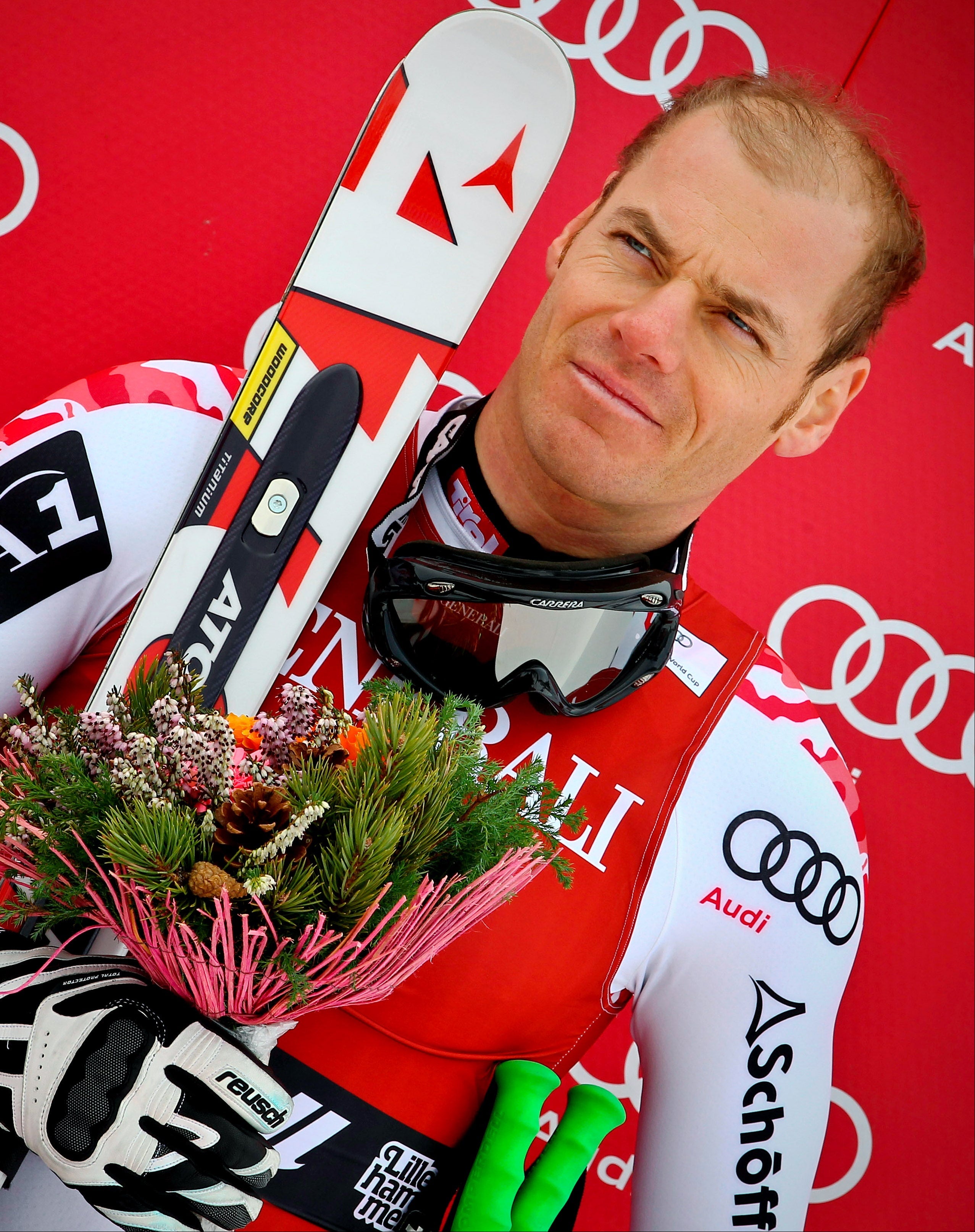 A close-up of ski racer Michael Walchhofer holding a bouquet of flowers and skis against a red sponsor backdrop. He is wearing a white and red ski racing suit with black and white gloves.