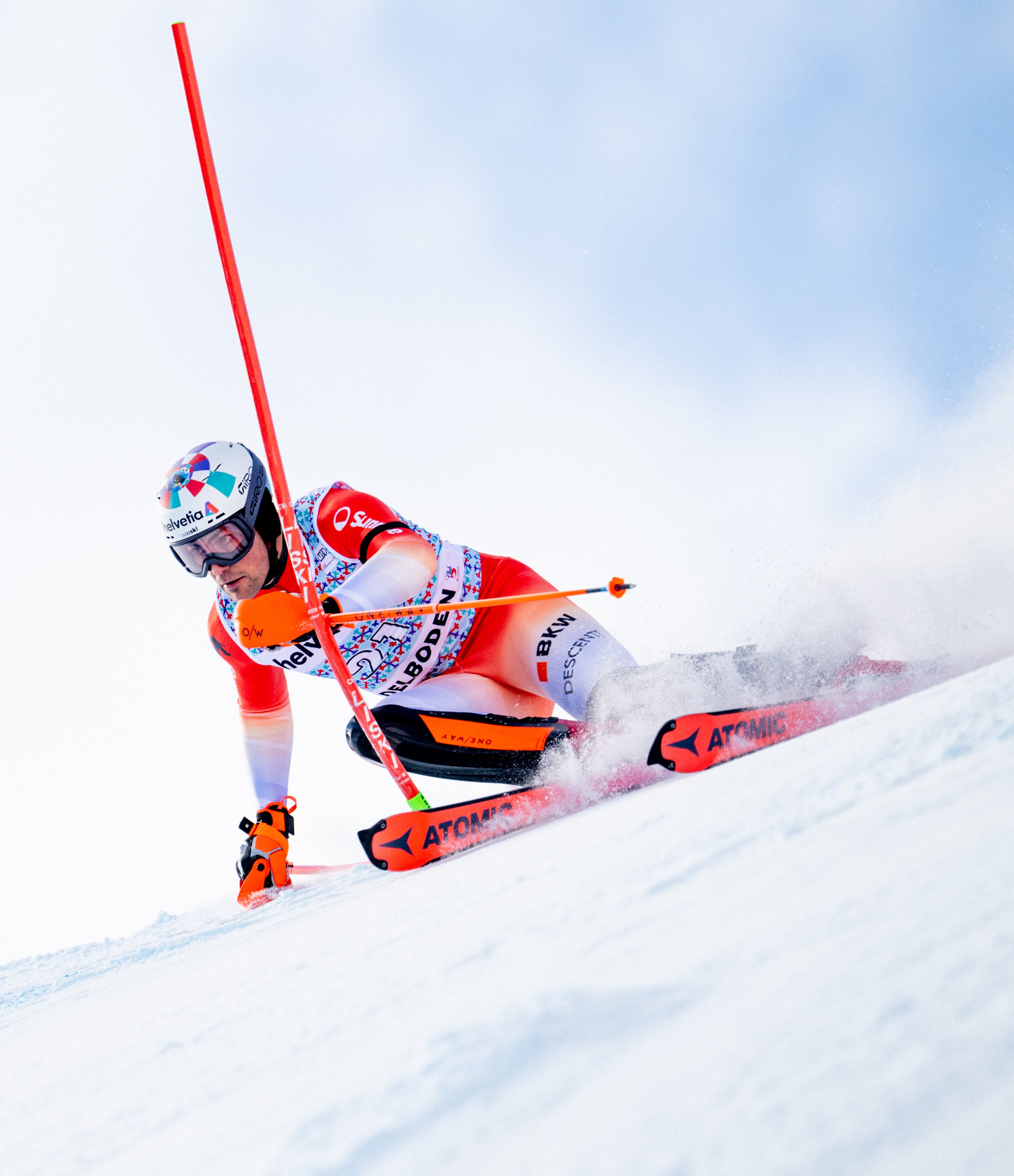 Daniel Yule in a red, white, and black racing suit leans sharply into a turn on a snowy slope, holding orange poles and skis with visible spray behind them. The skier wears a patterned helmet and goggles, and the background is a bright snowy sky.