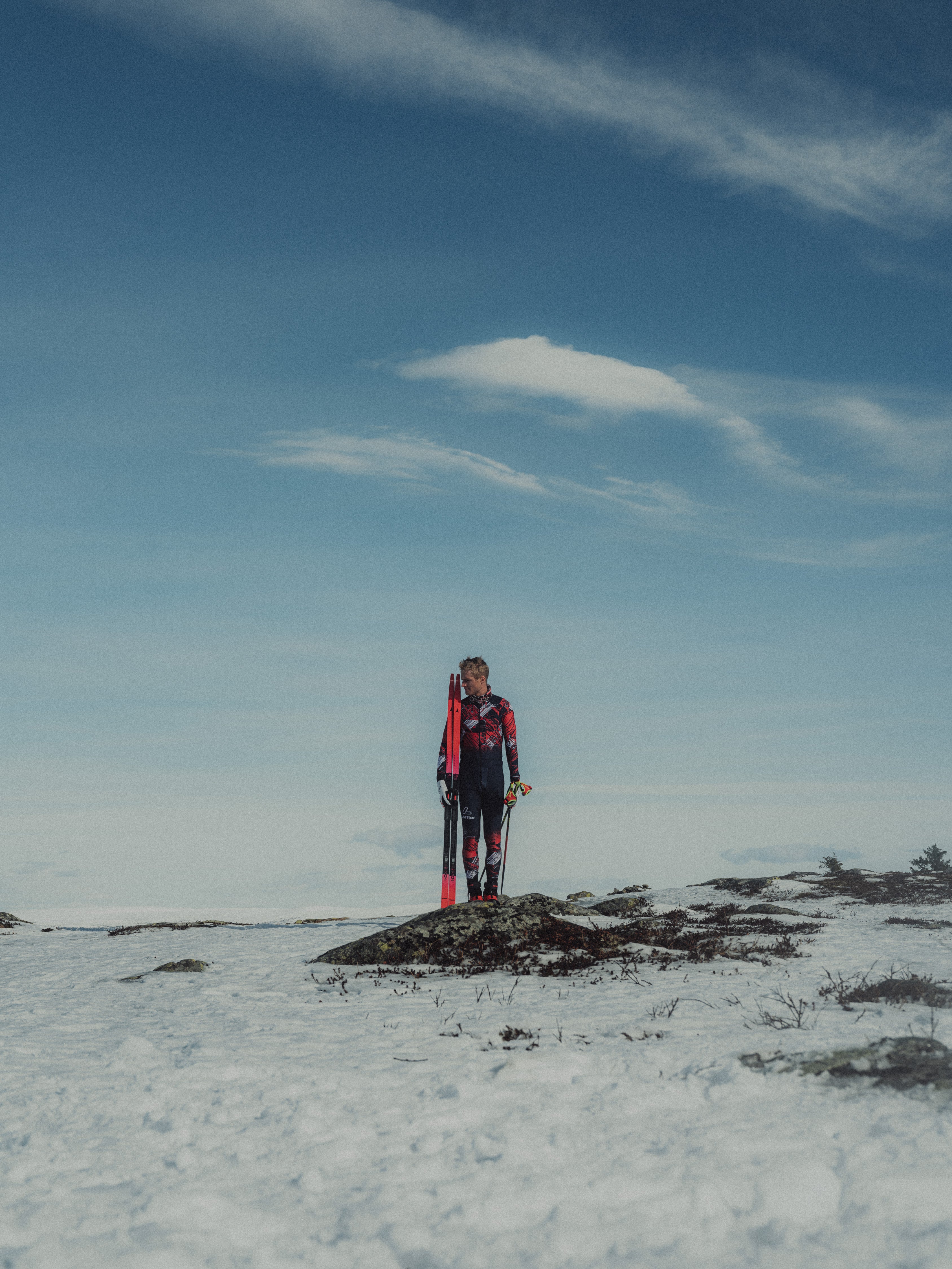 Mika Vermeulen standing outdoors in a snowy landscape on a small rocky patch, wearing a dark ski suit and holding ski poles, with skis visible behind them under a blue sky with light clouds.