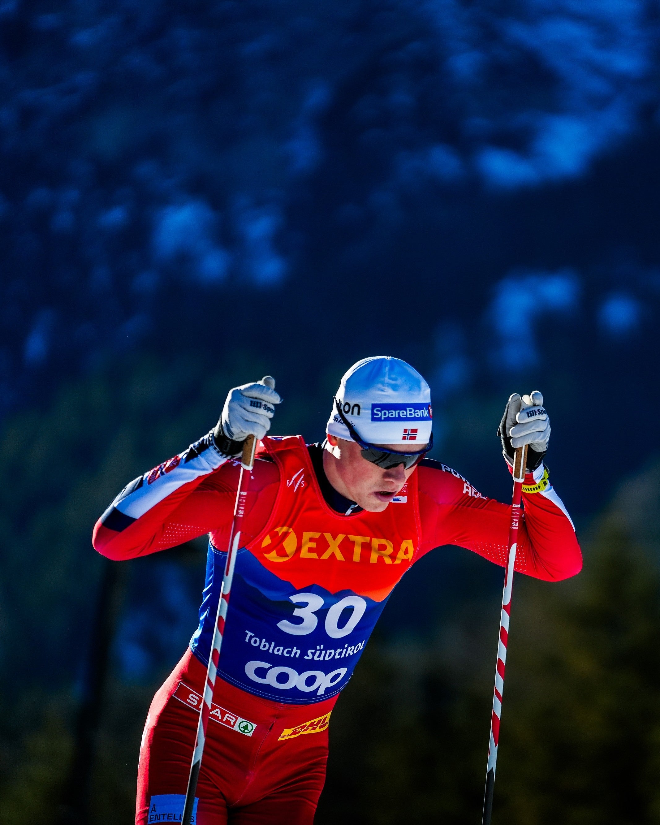 A cross-country skier in a red and blue ski suit with 'EXTRA' and 'COOP' branding, wearing a blue helmet, captured mid-action against a dark blue mountain background.