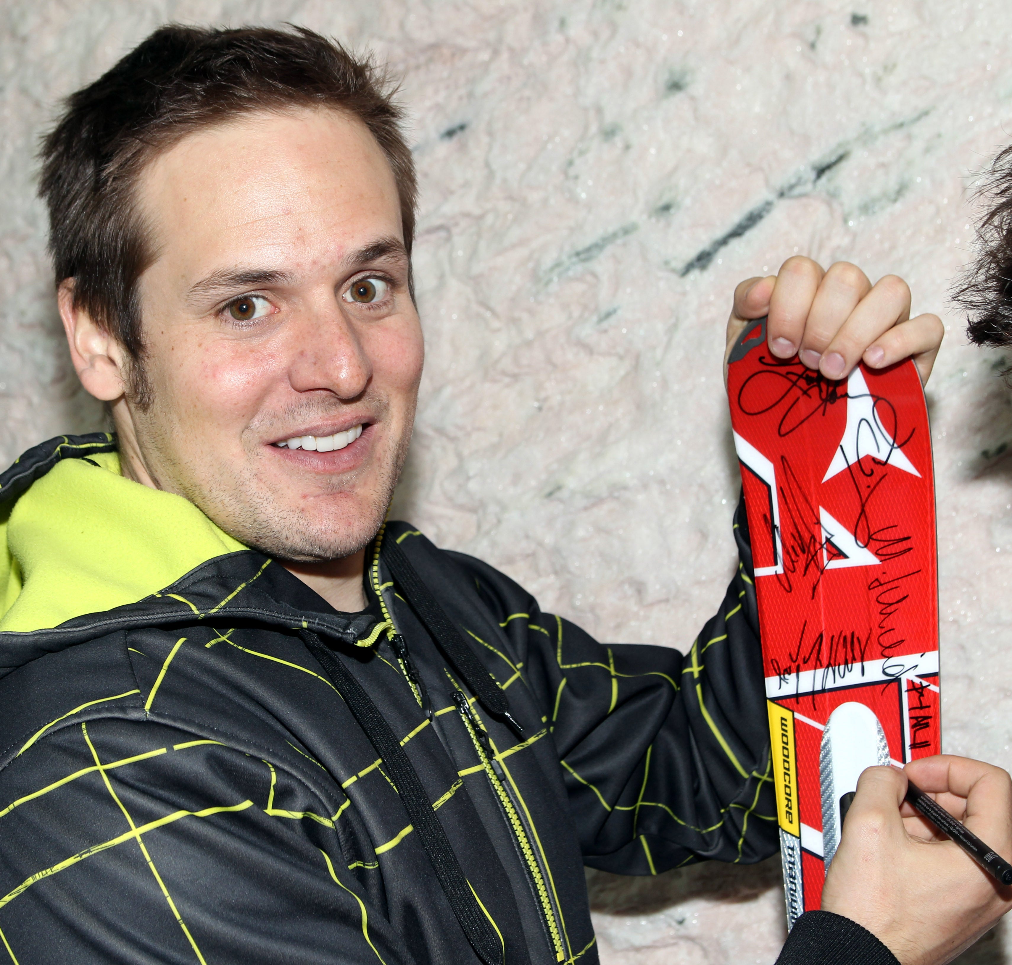 Daniel Albrecht in a black and neon yellow zip-up jacket signs a red and white autographed ski board with a black marker.