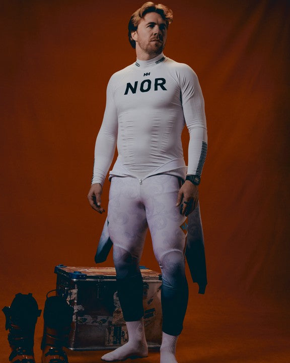 Aleksander Aamodt Kilde stands against a warm orange-brown backdrop wearing a fitted white long-sleeve athletic baselayer with 'NOR' printed across the chest and matching light gray patterned leggings. He is barefoot with white socks, and dark gear sits on the floor beside him.