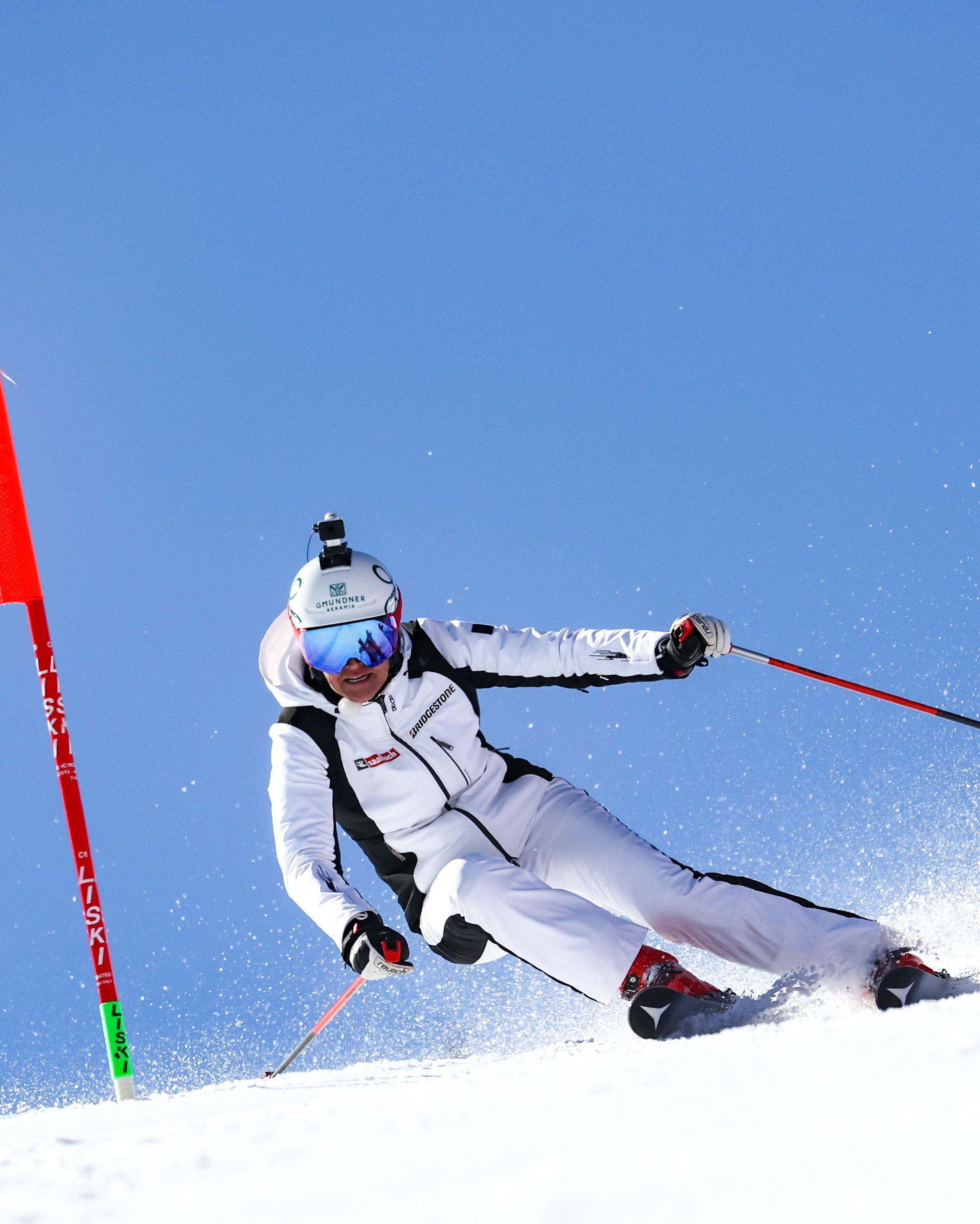 Alexandra Meissnitzer in a white racing suit with black trim and blue goggles leans sharply into a turn on a snowy slope, passing a red ZGONC gate flag under a clear blue sky.