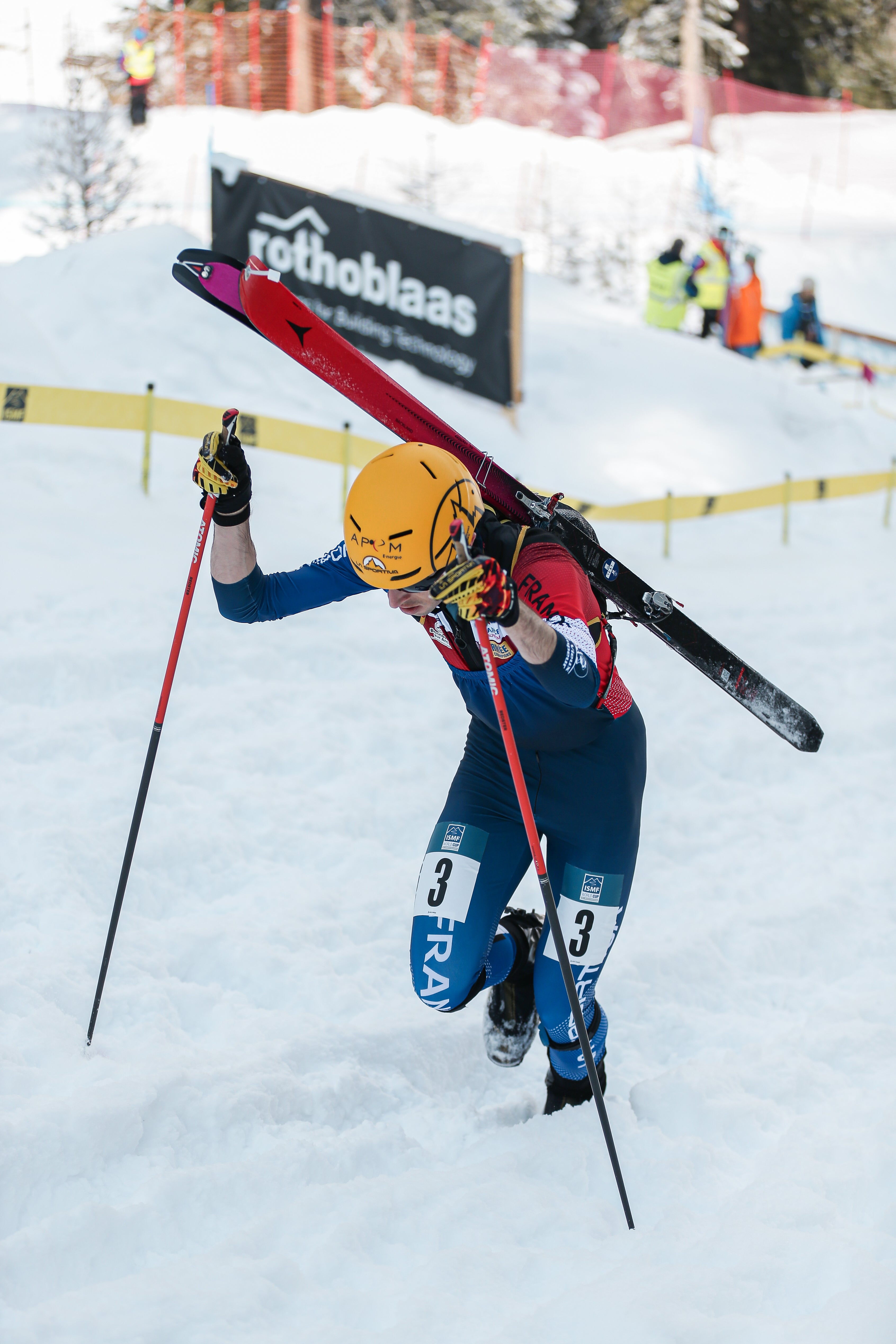Thibault Aselmet in a blue and red suit jumps climbs uphill carrying red skis on his back, using ski poles for support in a snowy outdoor setting.