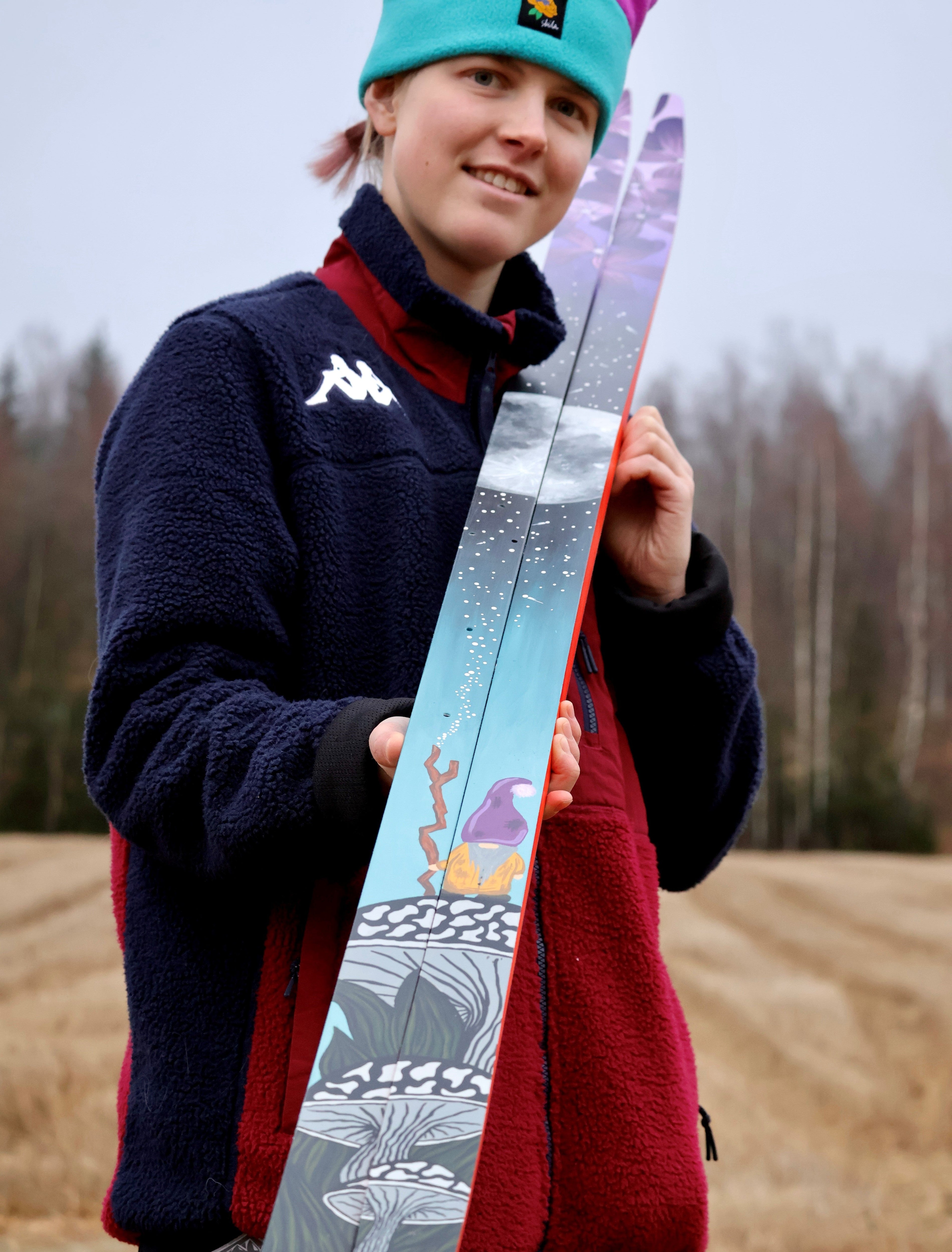 Alexa Brabec outdoors in a field wearing a navy fleece jacket with a red inner layer and holding a pair of nordic skis with a colorful winter graphic design.