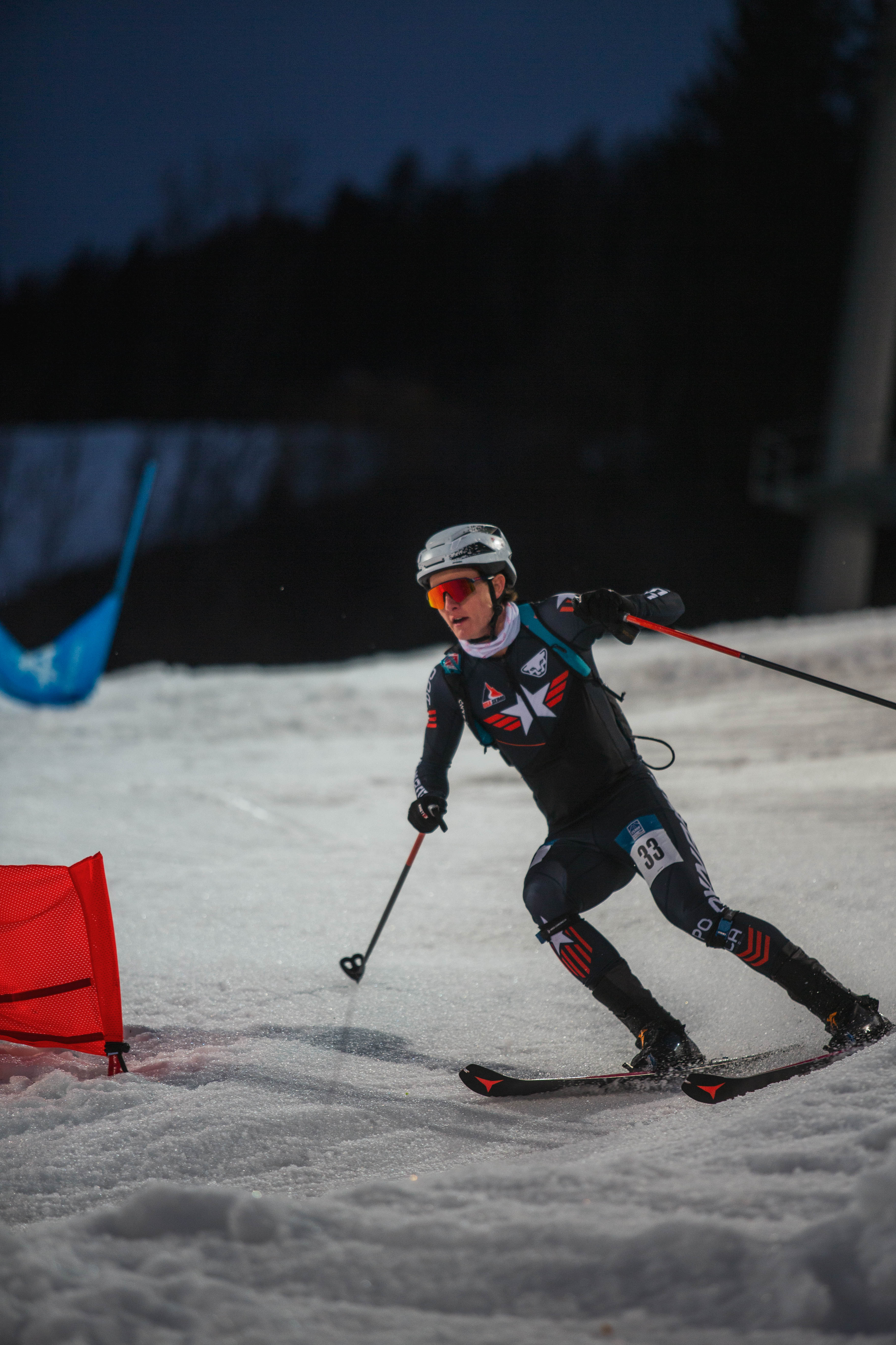 Griffin Briley in a black racing suit with red, white, and blue graphics, orange sunglasses, and a white helmet gliding through snow during a race.