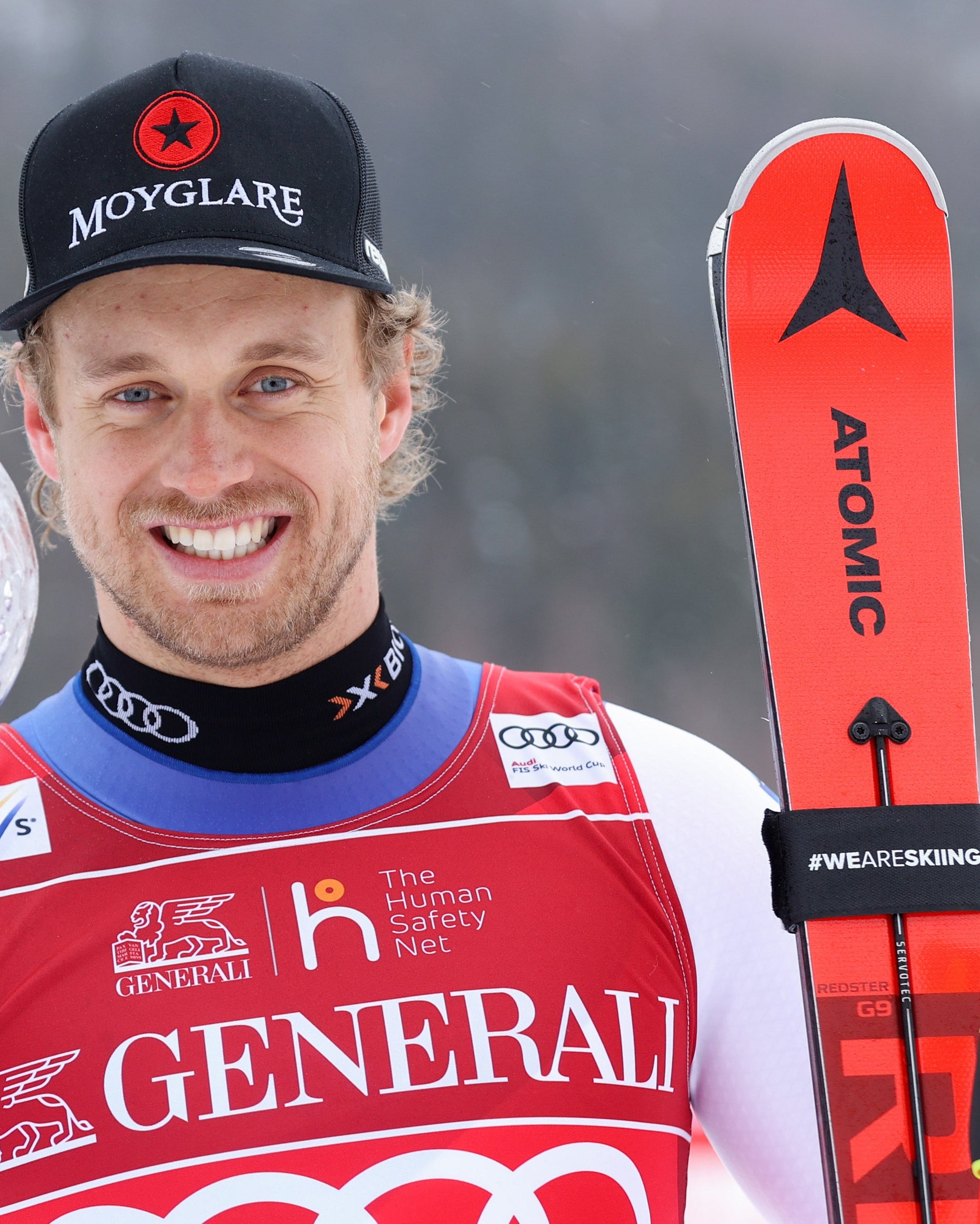 A smily Mauro Caviezel  in a red and white racing suit holds a large clear trophy cup and stands beside red skis. He wears a black cap, photographed outdoors in a snowy setting.