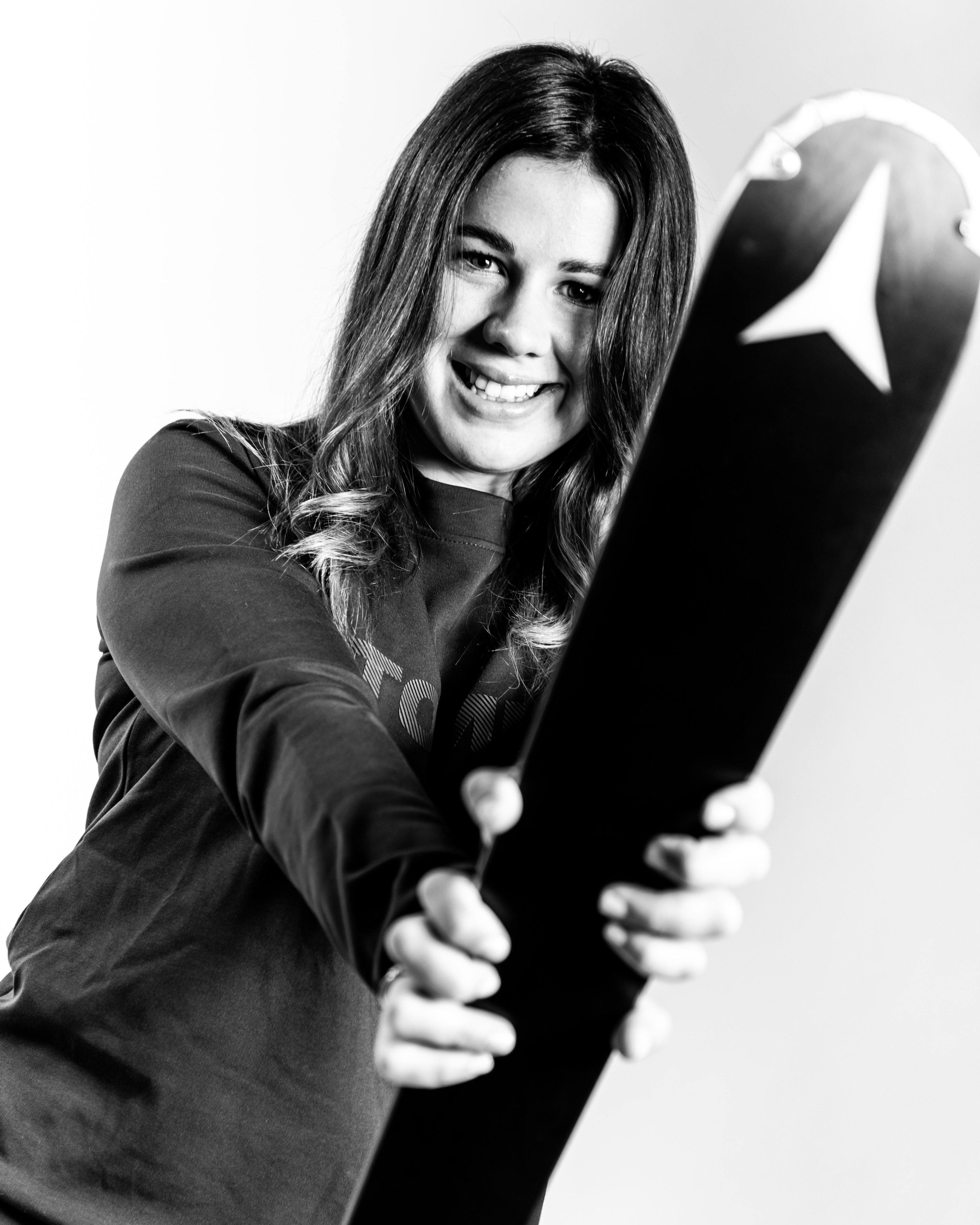 Nadia Delago holding a ski with a white Atomic logo on the sole, photographed in black and white against a light studio background.
