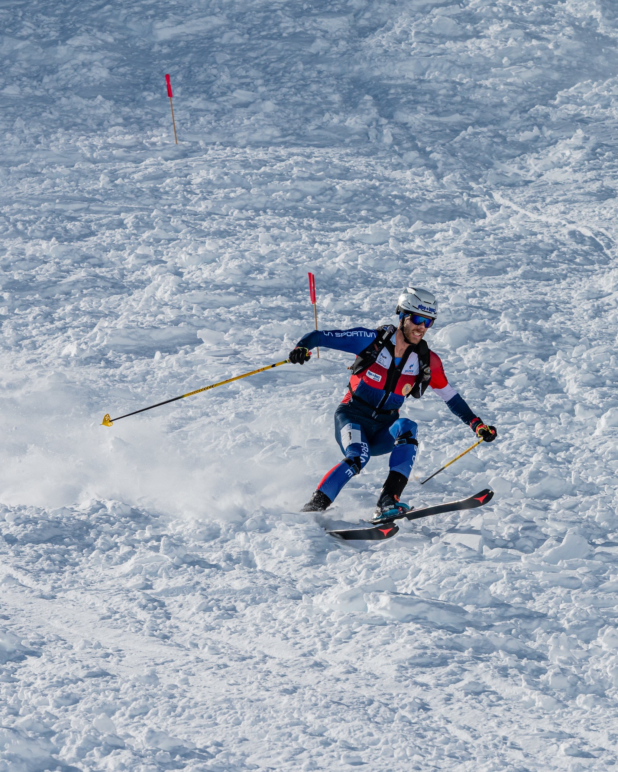 Samuel Equy in a blue, red, and black racing suit with a helmet and poles carving through deep snow on a snowy mountain slope.