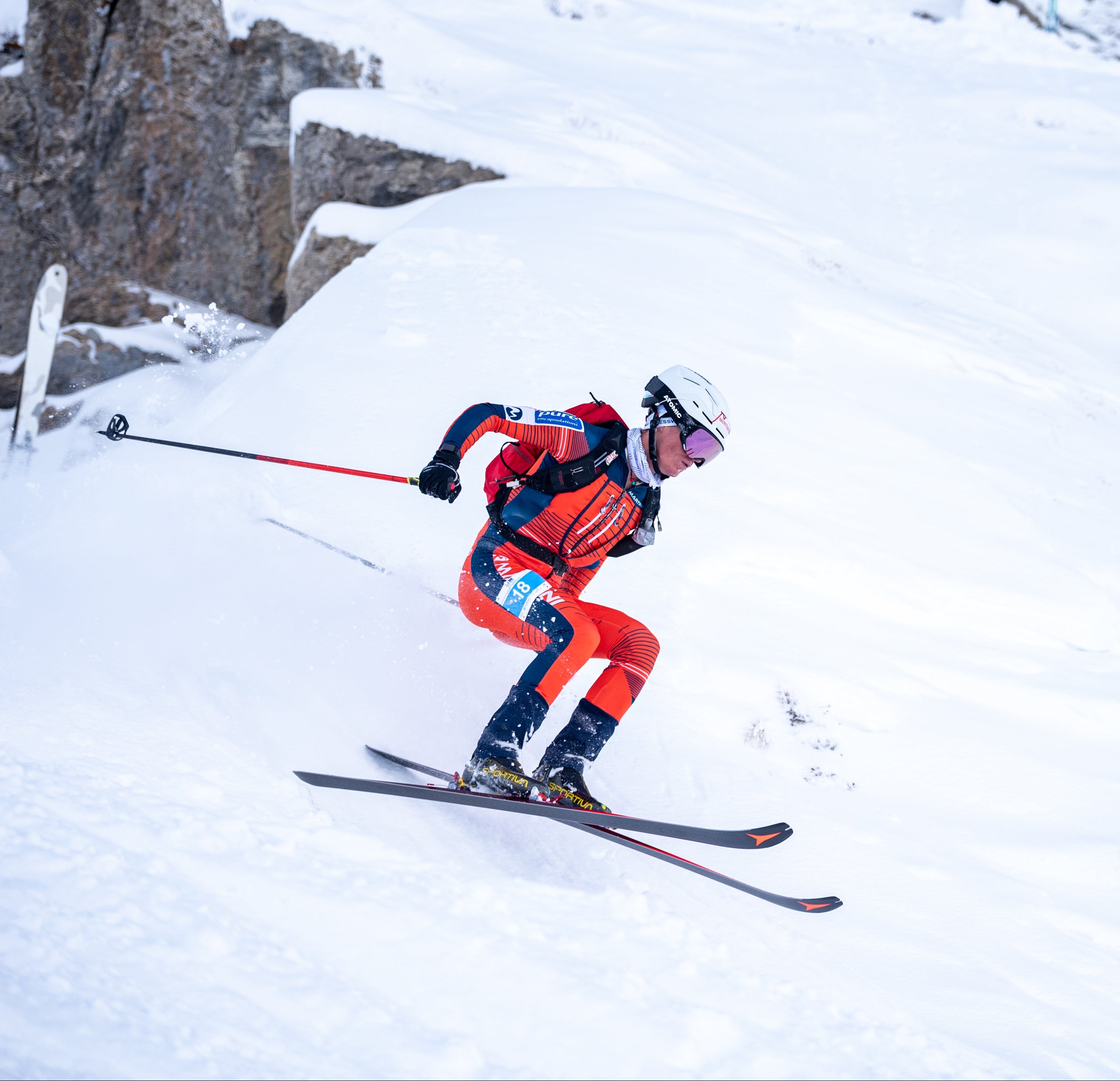 Daniel Ganahl in a red, navy, and white racing suit glides through deep snow, holding ski poles and wearing a white helmet and goggles.