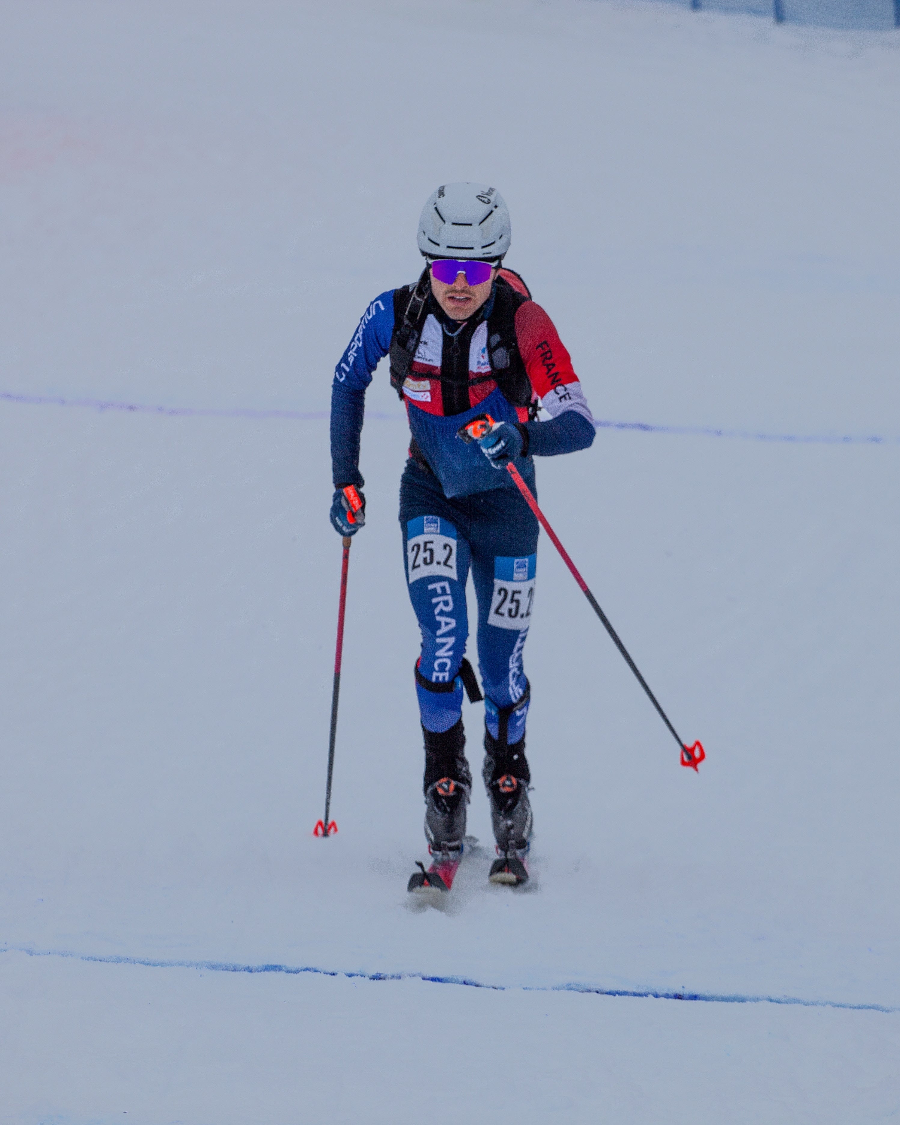 Pablo Giner wearing a blue, red, and black racing suit with 'FRANCE' on the pants, a white helmet, and purple-tinted sunglasses, gliding on a snowy track.