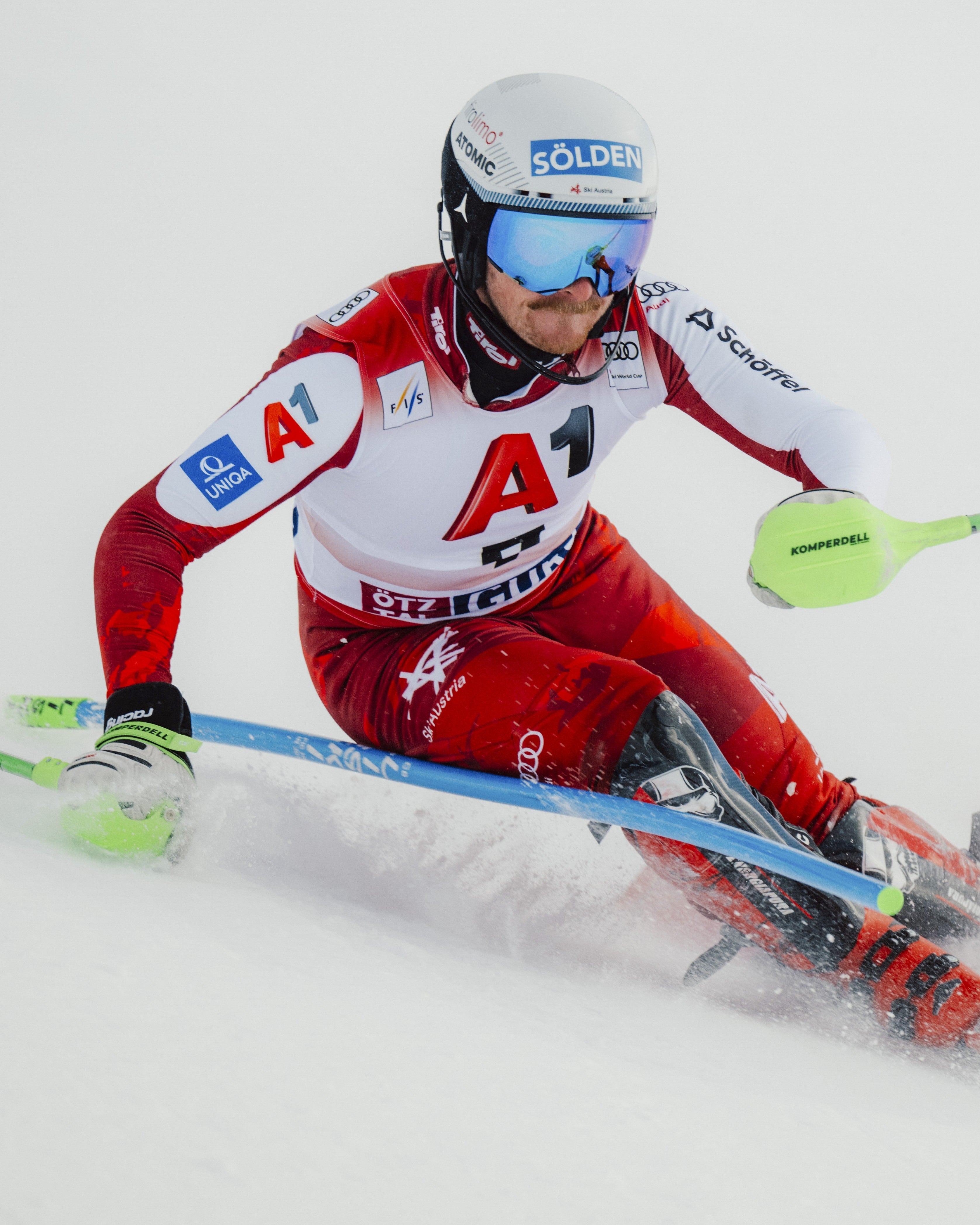 Fabio Gstrein in a red and white racing suit leans sharply through slalom gates, holding bright green ski poles. The skier wears a white helmet with blue goggles and visible sponsor logos.