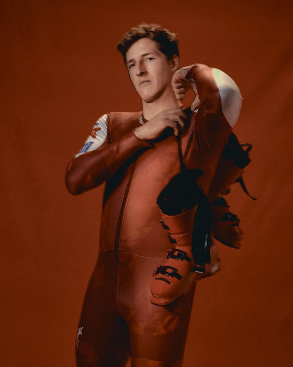 Raphael Haaser wearing a fitted orange-brown racing suit, holding a pair of ski boots against a warm orange-brown studio backdrop.