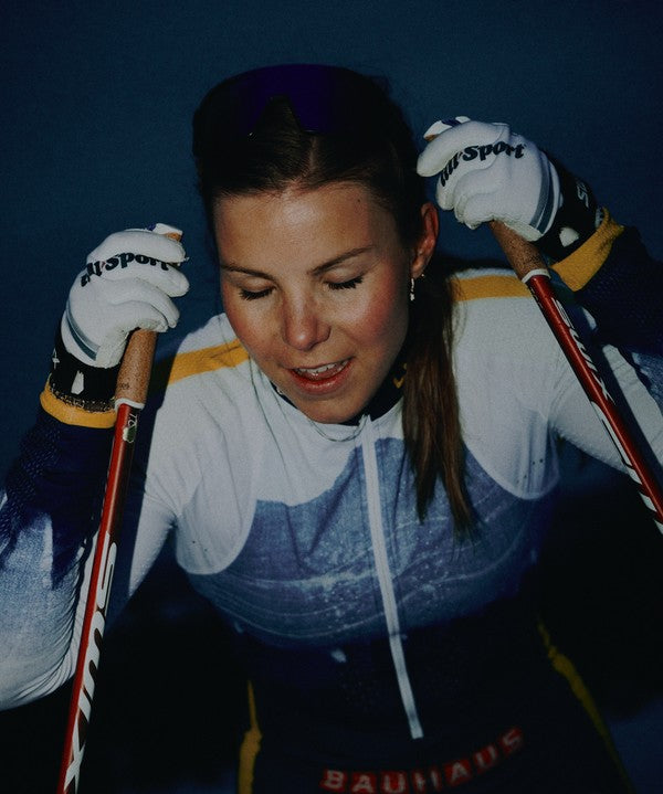 Johanna Hagstroem in a white, blue, and yellow sracing suit with white gloves, holding cross country skiing poles against a dark blue background.