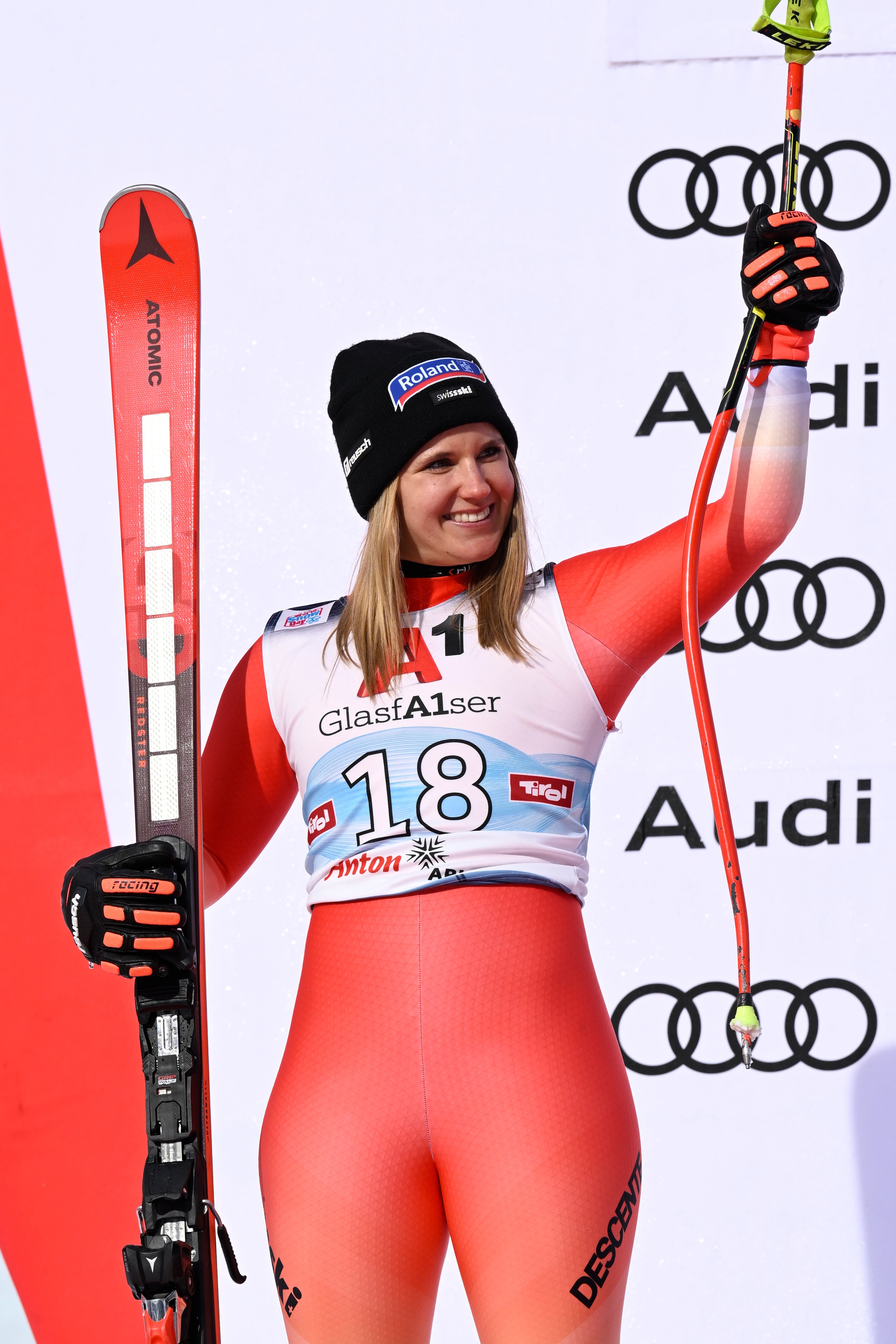 Joana Hählen standing on a podium, holding a red ski in one hand and ski poles in the other, wearing a bright red and white competition suit with the number 18 and sponsor logos including.