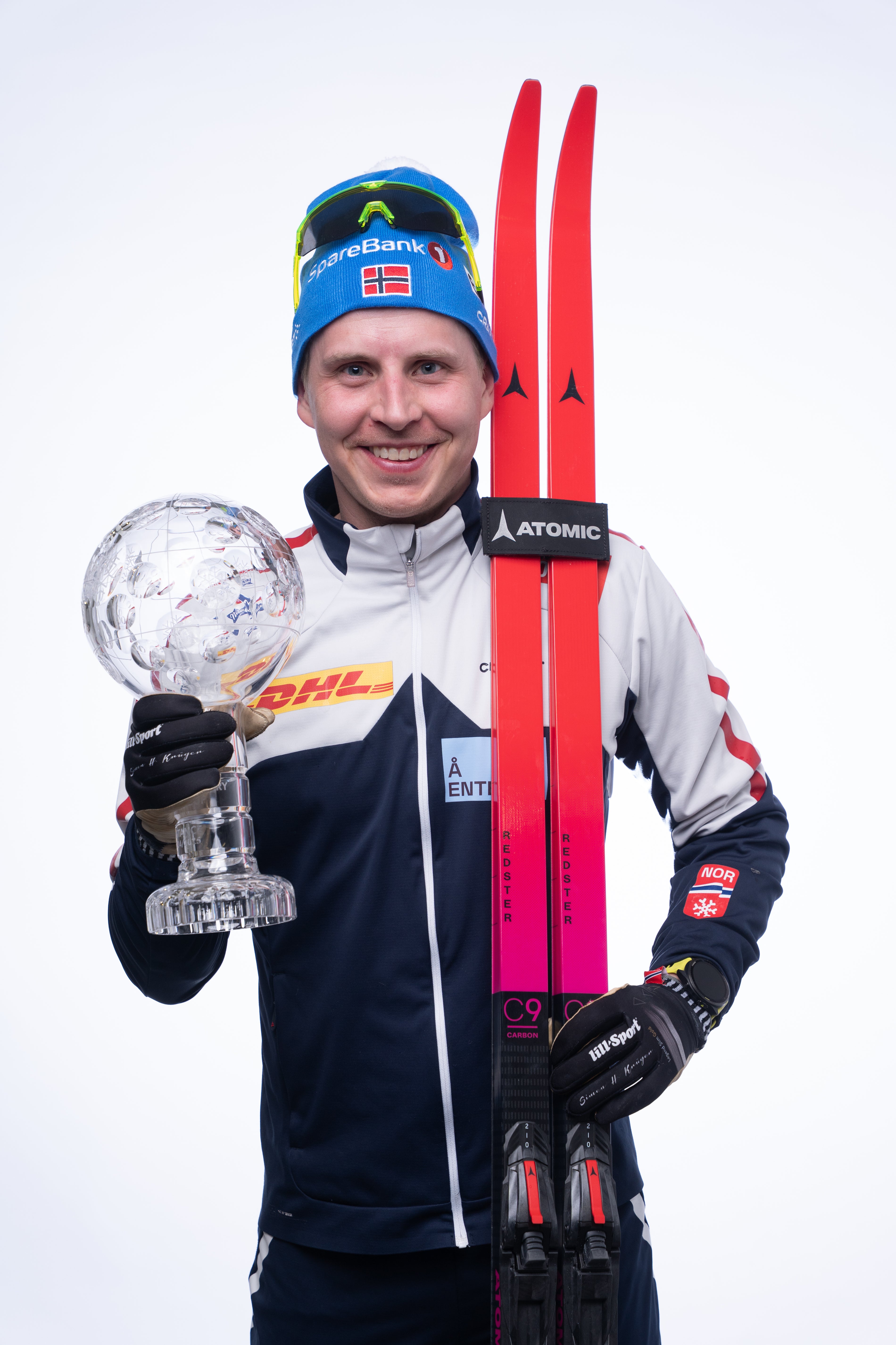 Simen Hegstad Krüger wearing a blue and white jacket, blue beanie, and black gloves, holding a clear crystal trophy and standing with red and pink skis against a white studio background.
