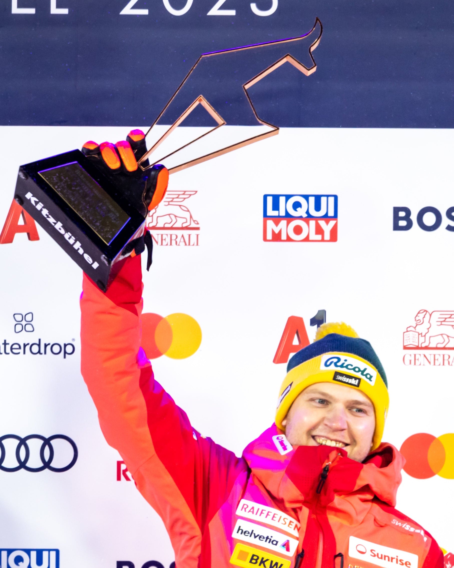 Niels Hintermann celebrates on a podium holding a trophy, wearing a bright red ski jacket, a yellow and blue beanie, and gloves. The background features sponsor logos including Liqui Moly, Audi, and Mastercard.