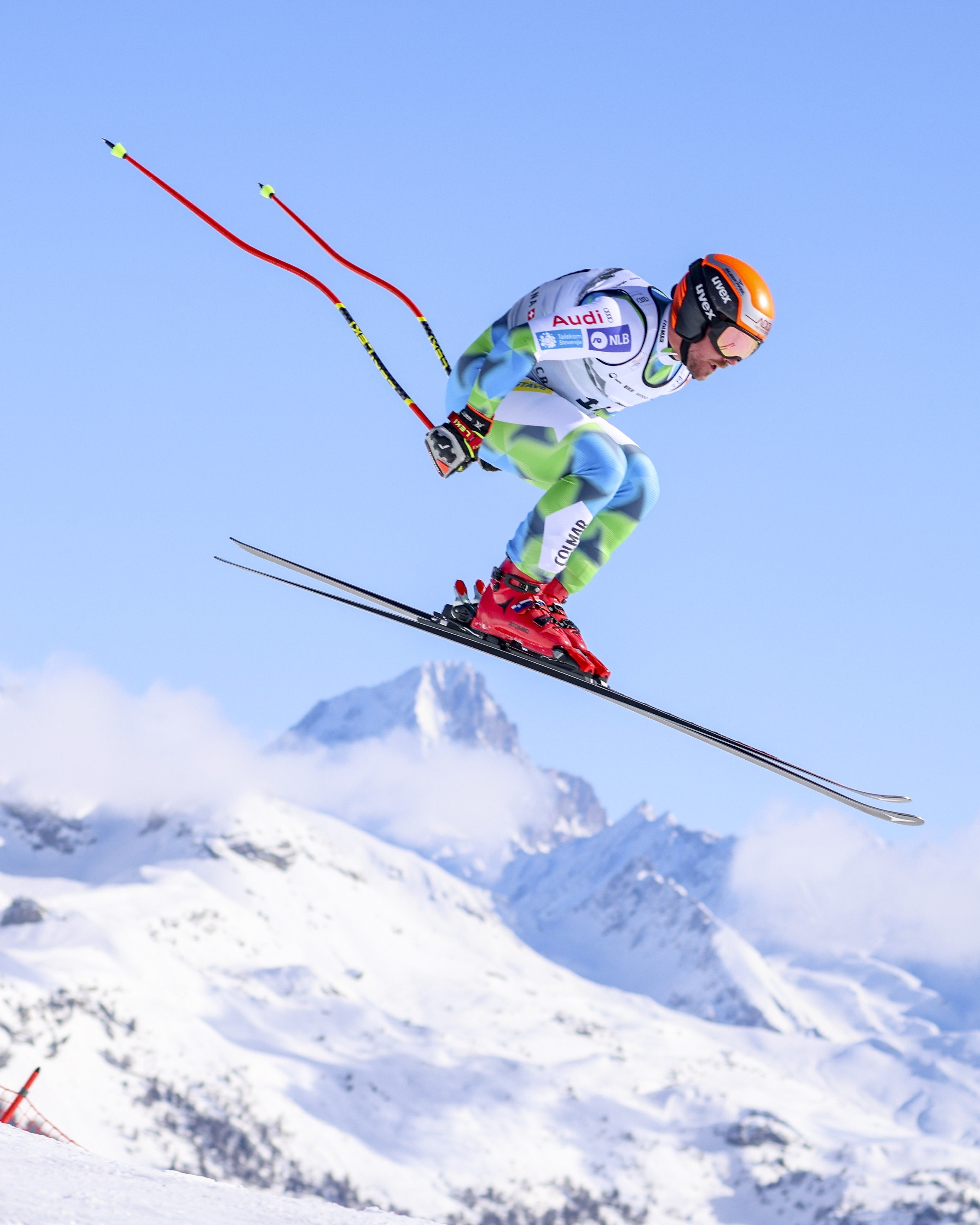 Miha Hrobat in a white, green, and blue racing suit with red boots and an orange helmet jumps high in the air during a speed race against a backdrop of snowy mountains and a clear blue sky.